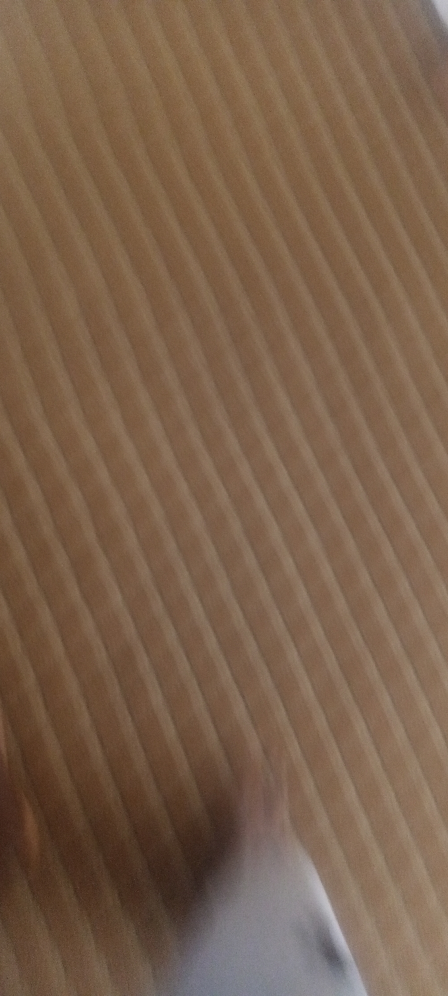 The image shows a close-up of a corrugated cardboard surface. The corrugations are oriented diagonally across the frame, with a brown hue. At the bottom of the image, a blurry, white object is partially visible. The location is unknown.