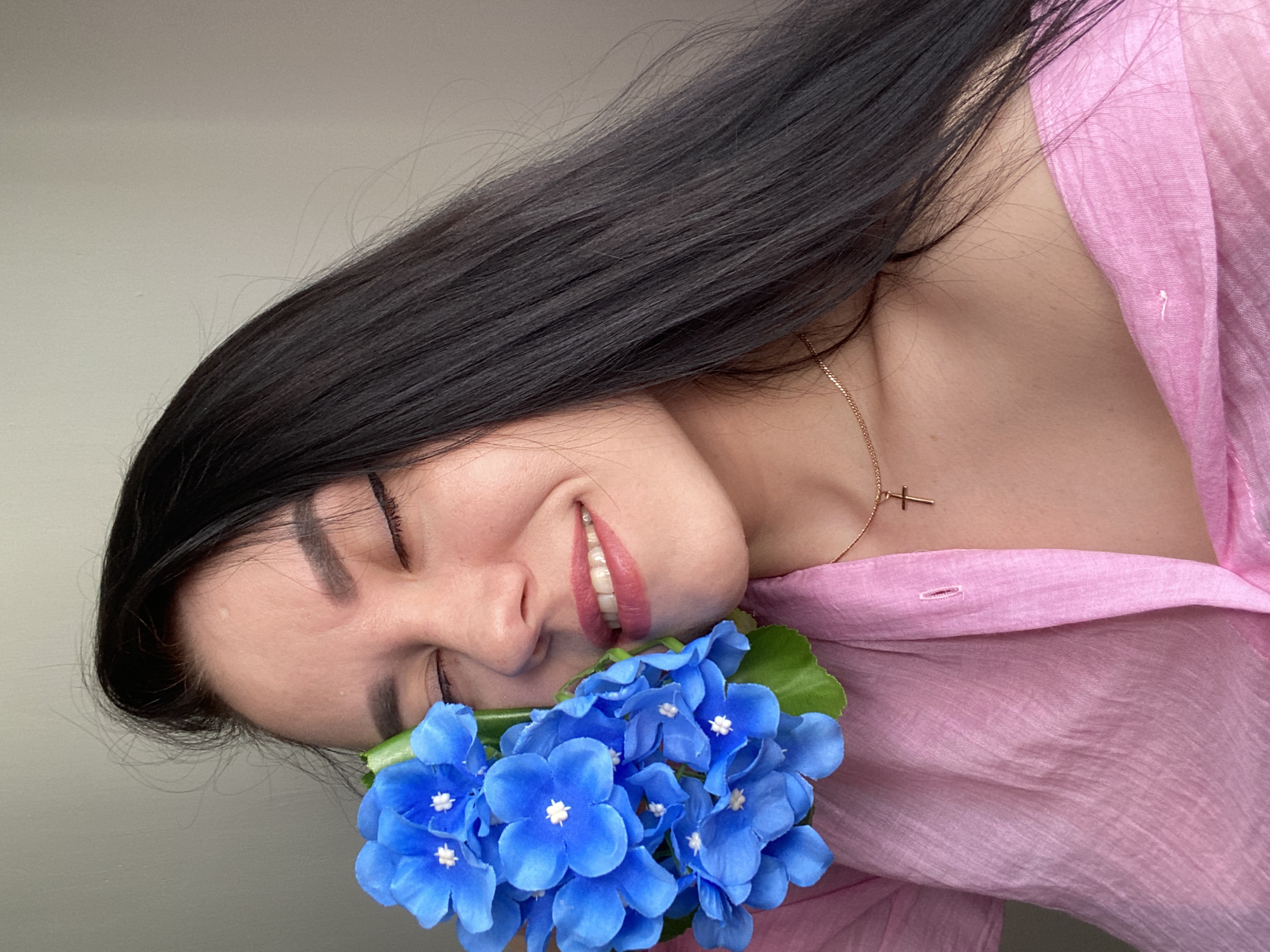 A woman is posing with her eyes closed and a smile on her face. She is holding a bouquet of bright blue flowers near her face. She has long, dark hair and is wearing a pink, button-down shirt. A gold necklace with a cross pendant is visible around her neck. The background is a neutral gray wall. The image was taken in a location with indoor lighting. There are no indicators of the city of Chaikovsky, Russia in the image.