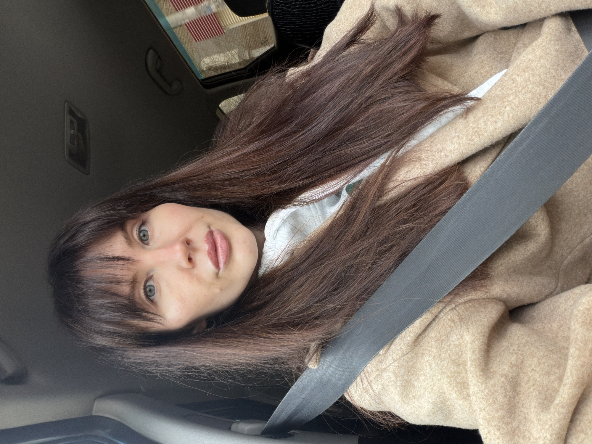 A woman with long brown hair and blue eyes is wearing a beige coat and a gray seatbelt in a car. The location is Perm, Russia, as indicated by the license plate or other visual clues not readily apparent in the image. The woman is looking at the camera. The car's interior is visible, including the headliner, a grab handle, and a portion of the dashboard. There is a sunroof open.