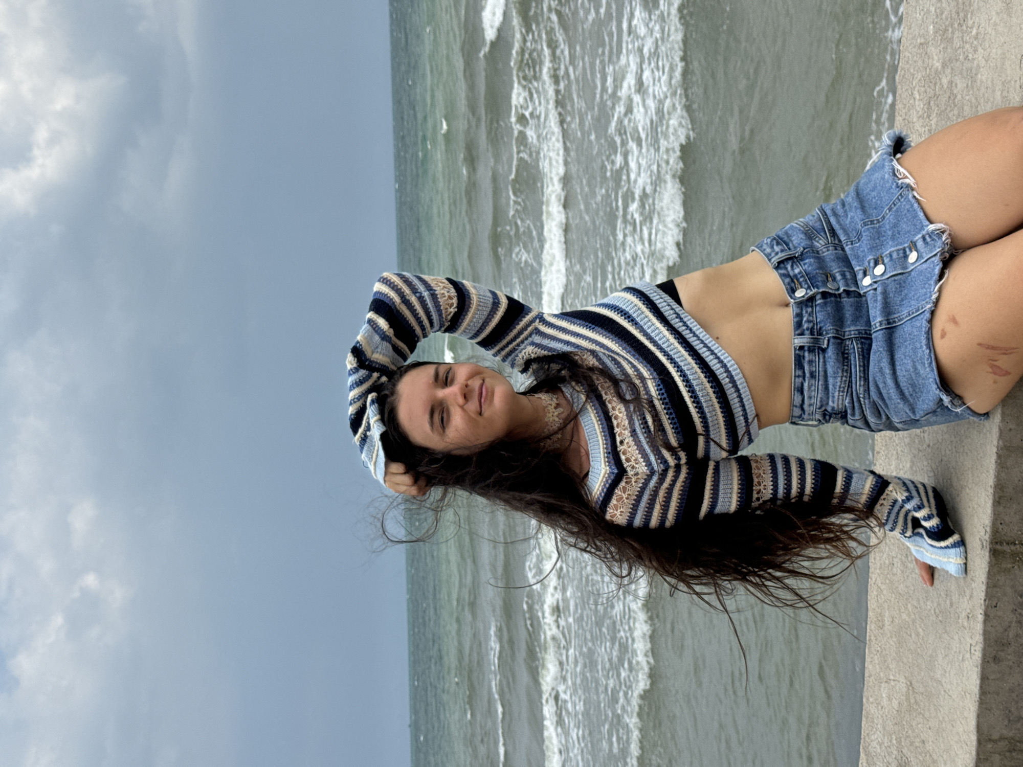 A young woman with long, dark hair is seated on a concrete wall overlooking a choppy, grey ocean under an overcast sky. She is wearing a striped, cropped sweater in shades of blue, white, and beige, and a denim mini-skirt. Her right arm is raised, with her hand resting on her head. Her hair is windswept. The lower part of her right thigh has visible abrasions and discoloration. The location is identified as Phu Thuy Ward, Vietnam.
