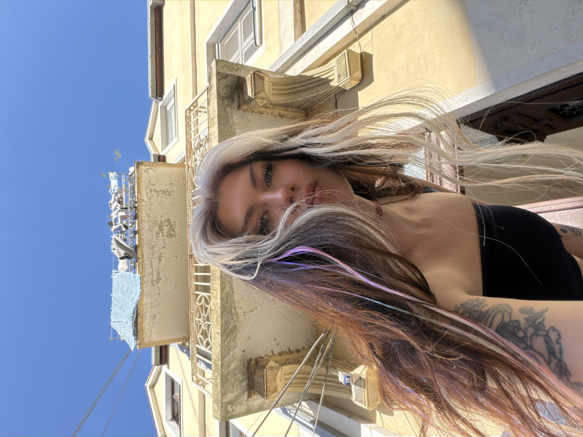 A portrait of a woman with light blonde and brown hair is angled upward. The woman's face and upper torso are the primary focus. She has fair skin, green eyes, and full lips. Her hair is long, with streaks of holographic color, and appears to be blowing in the wind. She wears a black top that reveals her shoulders. Tattoos are visible on her right arm.
Behind the woman is an old, yellow building with ornate architectural details, including balconies with metal railings. The sky is a clear blue.
Based on the architectural style and information about the woman's location provided with the image, this photo was likely taken in Belgrade, Serbia.