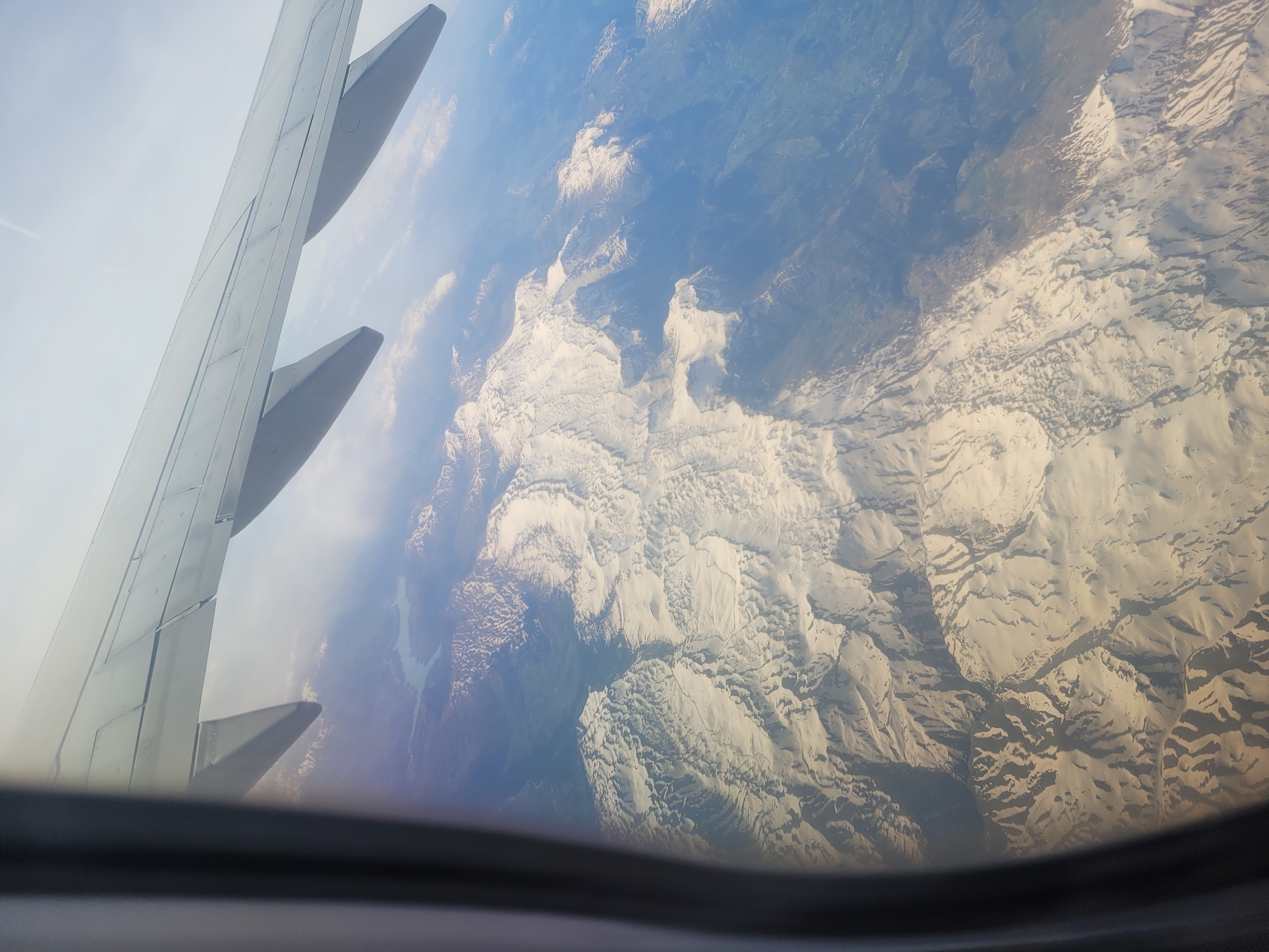 A view from an airplane window shows the aircraft's wing and flaps on the left and a snowy mountainous landscape below. The landscape features a mix of snow-covered peaks and valleys, with some areas appearing to be bare rock or vegetation. The sky is a pale blue. The airplane window frame is visible at the bottom of the image. The location in the city of Gündoğmuş, Turkey is unknown.