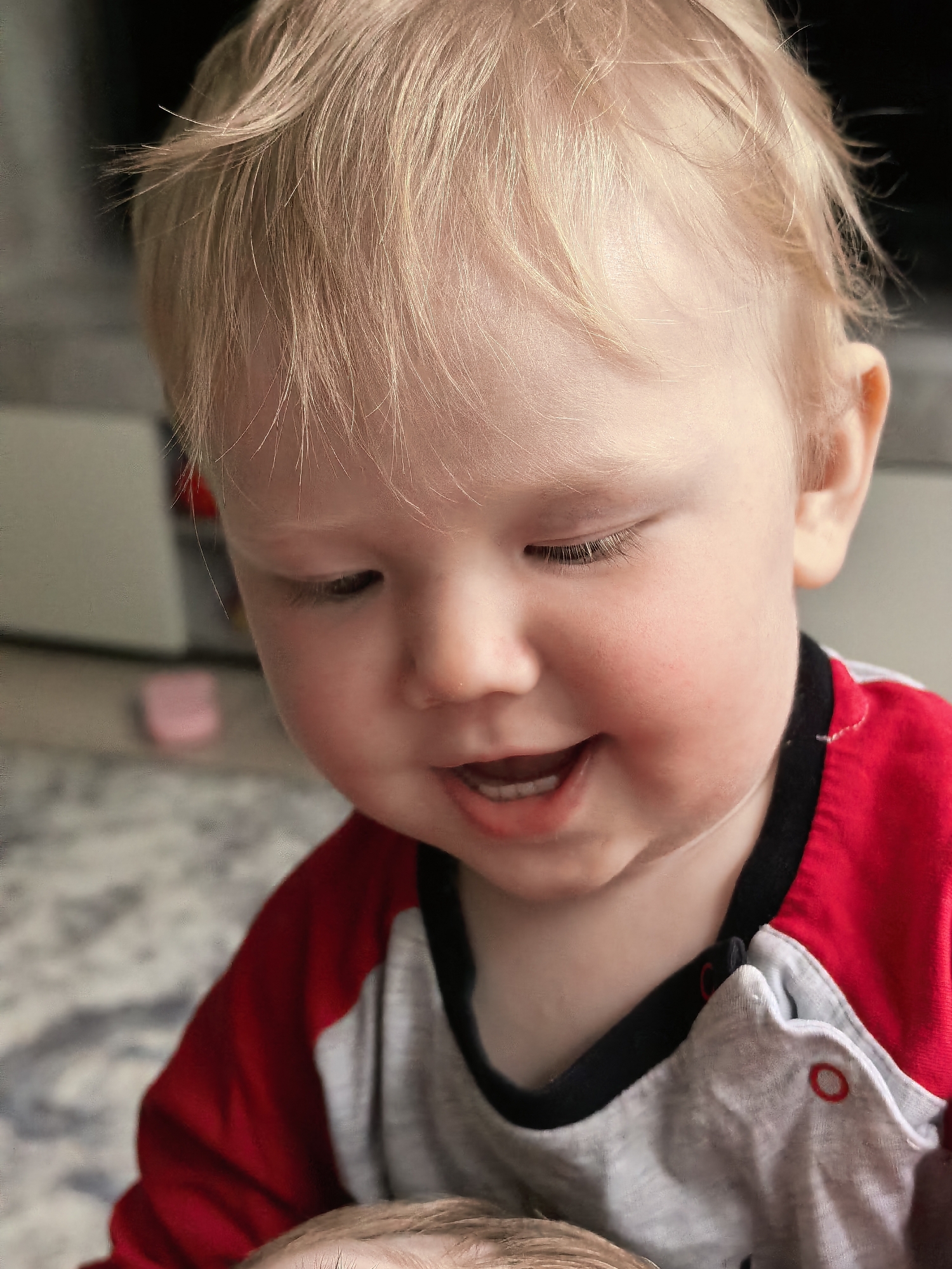 This is a close-up, waist-up, indoor photograph of a young toddler. The toddler has fair skin, rosy cheeks, and wispy blonde hair. They are looking down and to the left with their mouth open in a smile, revealing a few small teeth. Their eyes are partially closed, and their eyelashes are prominent.
The toddler is wearing a two-tone shirt with gray on the torso and red sleeves. The collar and cuffs are trimmed in black. The shirt appears to be a onesie, with visible red snap details on the front.
The background is softly blurred, suggesting an indoor setting. Hints of furniture and a patterned rug are visible. The lighting appears to be natural, possibly from a window, illuminating the toddler's face. The overall mood of the image is joyful and innocent, capturing a candid moment of a happy child. There is no visible text in the image.