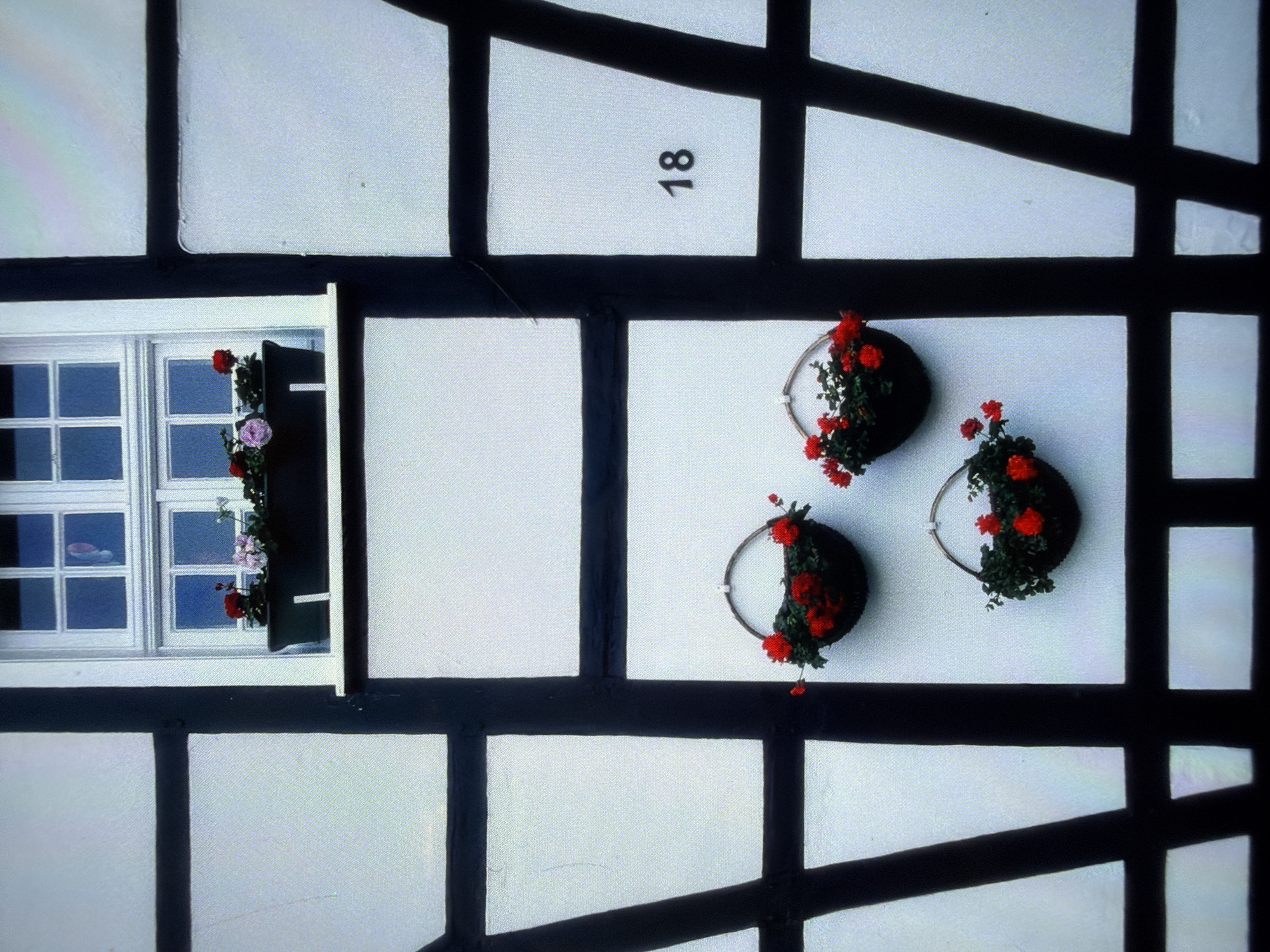 The image shows a close-up of the exterior of a building with a half-timbered architectural style. The facade is predominantly white with dark, thick beams forming a grid pattern. A white-framed window with multiple panes is visible in the upper section, with a window box filled with red and pink flowers situated below it. Hanging from the wall below the window are three circular baskets filled with red flowers and greenery. The number "18" is prominently displayed on one of the white sections of the facade, to the right of the baskets. The overall scene suggests an outdoor setting in front of a building.