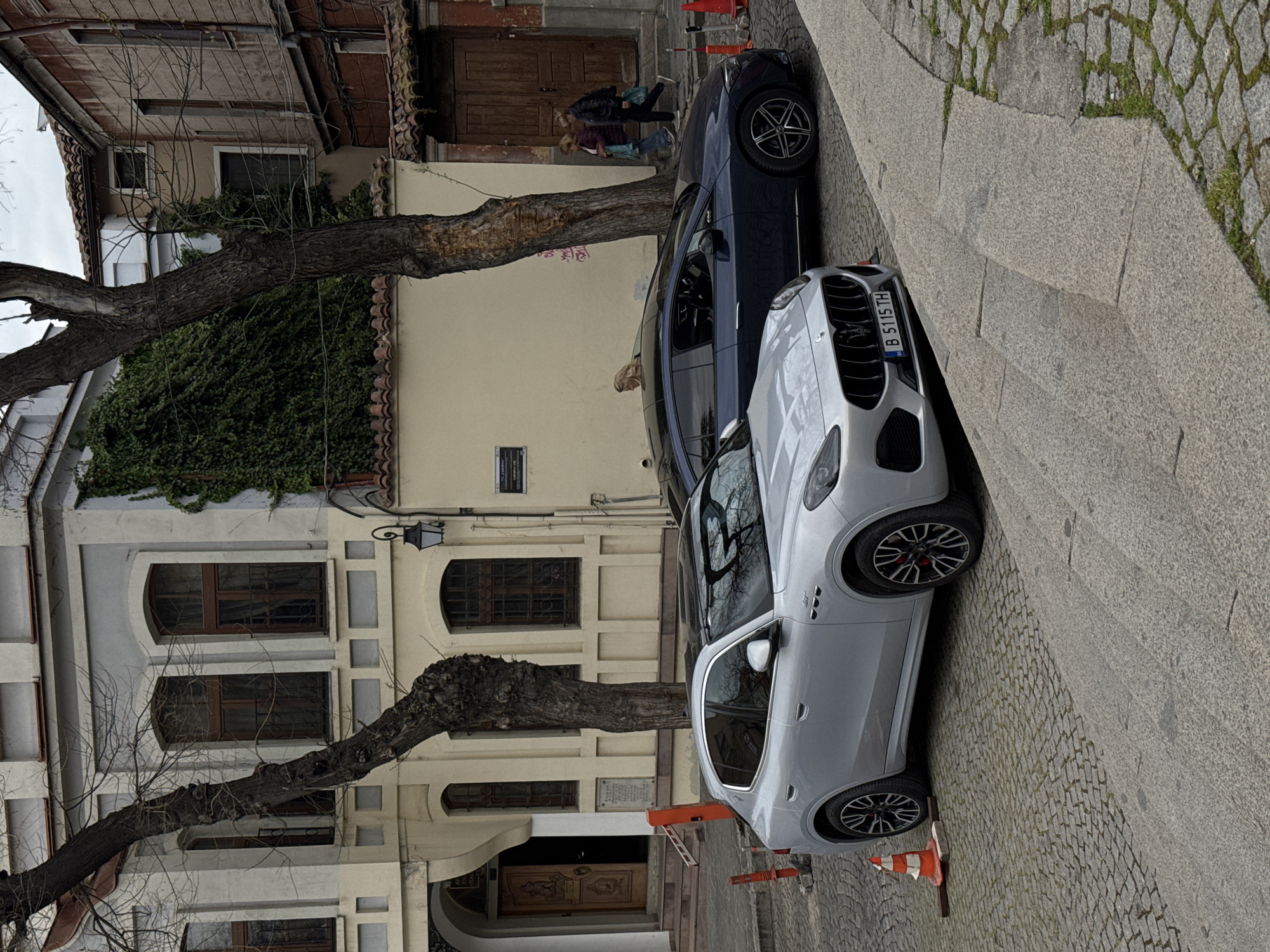 This street-level photograph captures a historical street scene in Plovdiv, Bulgaria, under an overcast sky. The perspective is slightly elevated, looking down a narrow cobblestone street lined with traditional buildings on the left and parked cars on the right.
In the foreground, a light grey Maserati Levante SUV is parked diagonally on the cobblestones, with its distinctive grille and emblem clearly visible. Its license plate reads "B 5115 TH," indicating a Bulgarian registration. Orange and white traffic cones are scattered on the street near the vehicle. Behind it, a dark blue car, possibly another SUV, is also parked.
On the left, old multi-story buildings showcase traditional architecture with arched windows, wooden frames, and security bars. One building is partly covered in green ivy, while another features a grand wooden entrance door. A vintage street lamp is mounted on a wall. Two large, mature trees with prominent, gnarled trunks and bare branches stand along the sidewalk, suggesting a cooler season.
Several people are visible near the buildings. Two women, one with blonde hair and one with dark hair, are standing near the wooden door, seemingly conversing. Another woman with blonde hair is partially visible behind the dark blue car.
A metallic plaque is affixed to one of the building walls. Partially decipherable text includes "МУЗЕЙ" (Museum), "ЕТНОГРАФСКИ" (Ethnographic), and "СТАРИНЕН ПЛОВДИВ" (Old Plovdiv), suggesting the location is an Ethnographic Museum within the historic part of Plovdiv. The scene depicts a calm, everyday moment in a culturally rich setting.