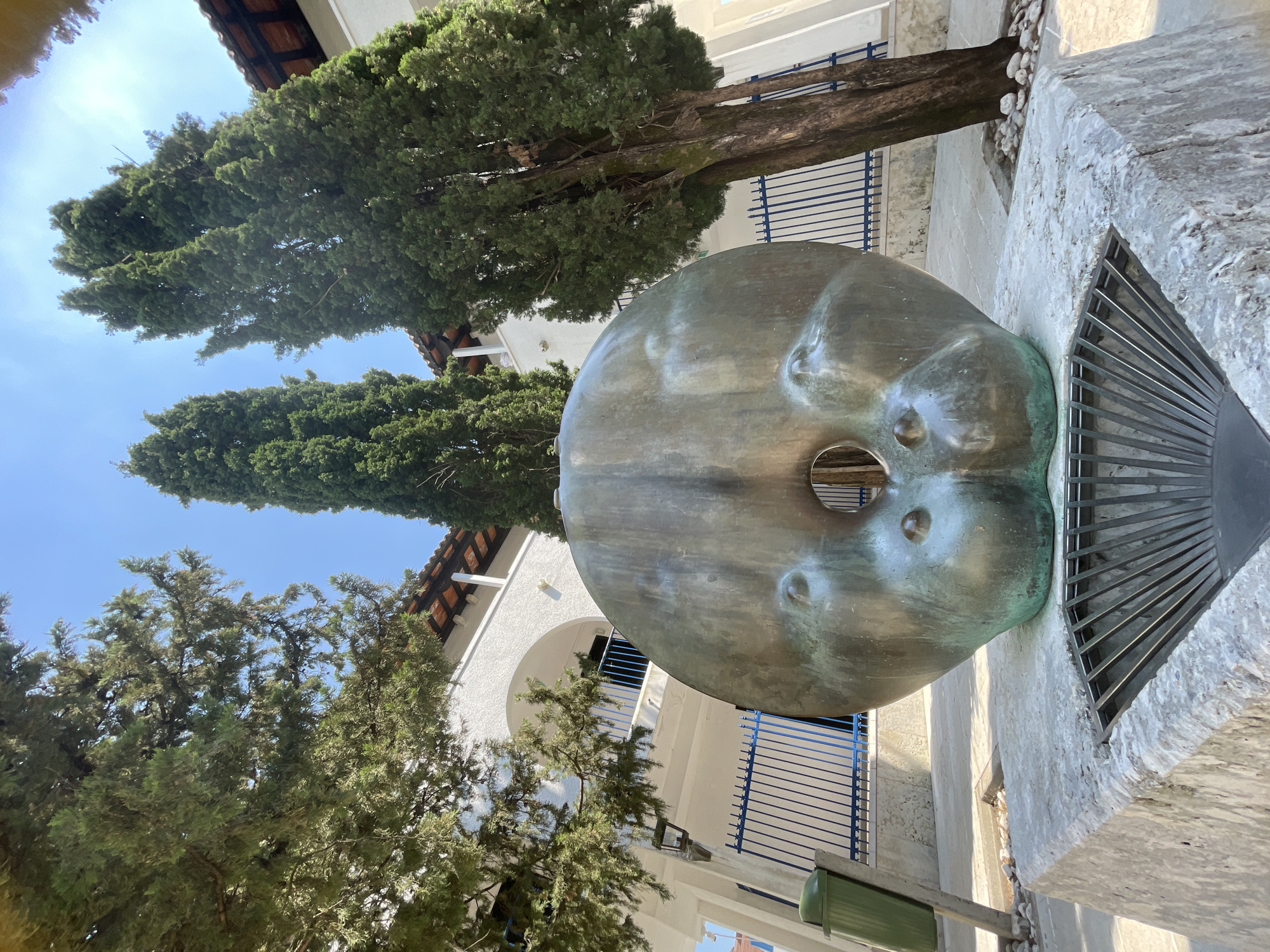 The image shows the "Head of a Woman" sculpture by Mitja Ribičič, located in Budva, Montenegro. The sculpture, a large bronze-colored head with a hole through it, rests on a stone base. The background includes trees, a building with white walls and a red-tiled roof, and blue railings. The sky is visible in the upper left corner. A green trash can is also visible in the lower right of the image. The image is shot from a low angle, looking up at the sculpture and the surrounding environment.