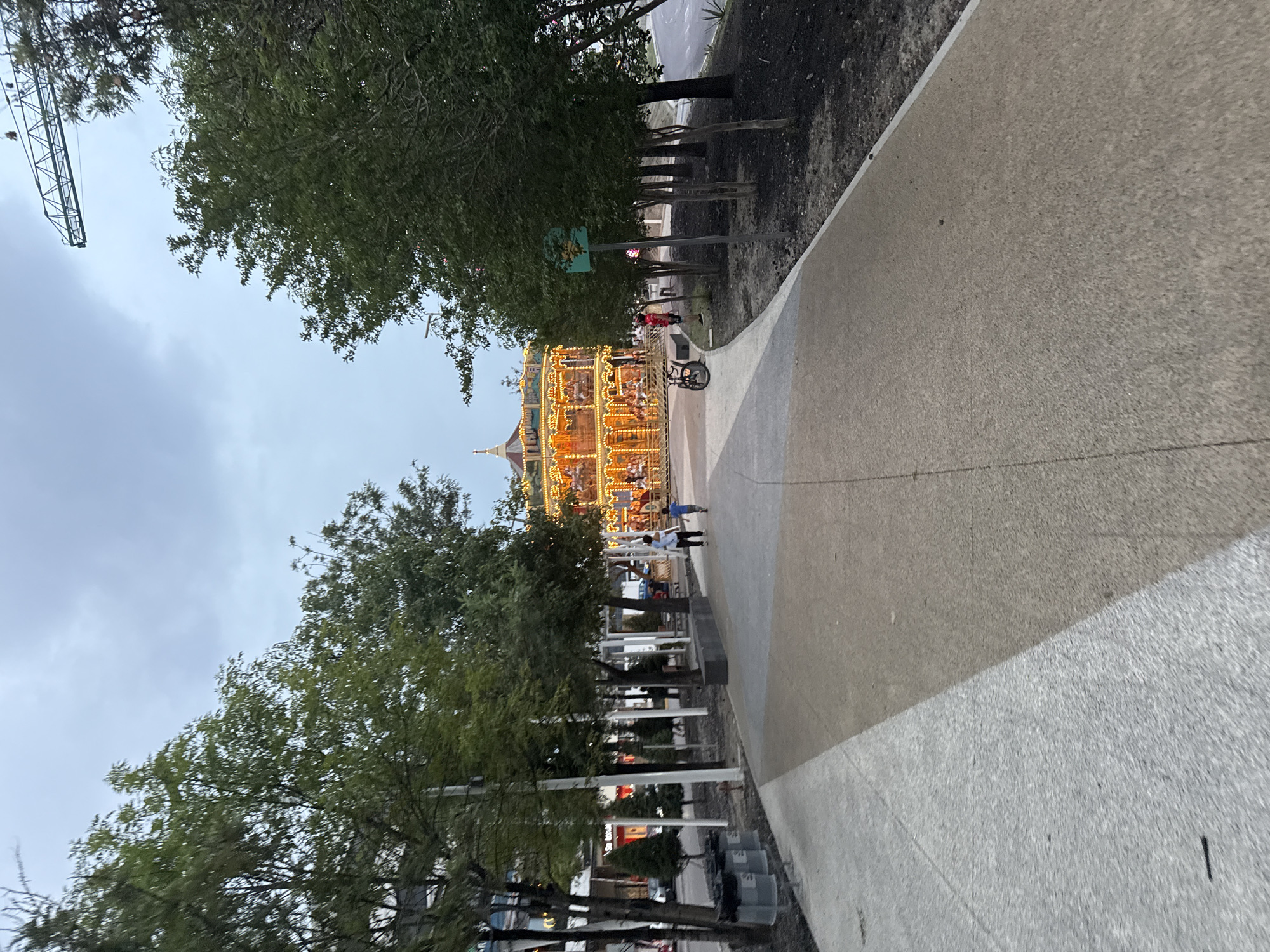 The image shows a carousel in Puebla, Mexico. The view is from a concrete pathway, with trees lining the sides. The sky is overcast. People are walking near the carousel. A bike is parked close to the carousel. A construction crane is visible in the top left of the image.