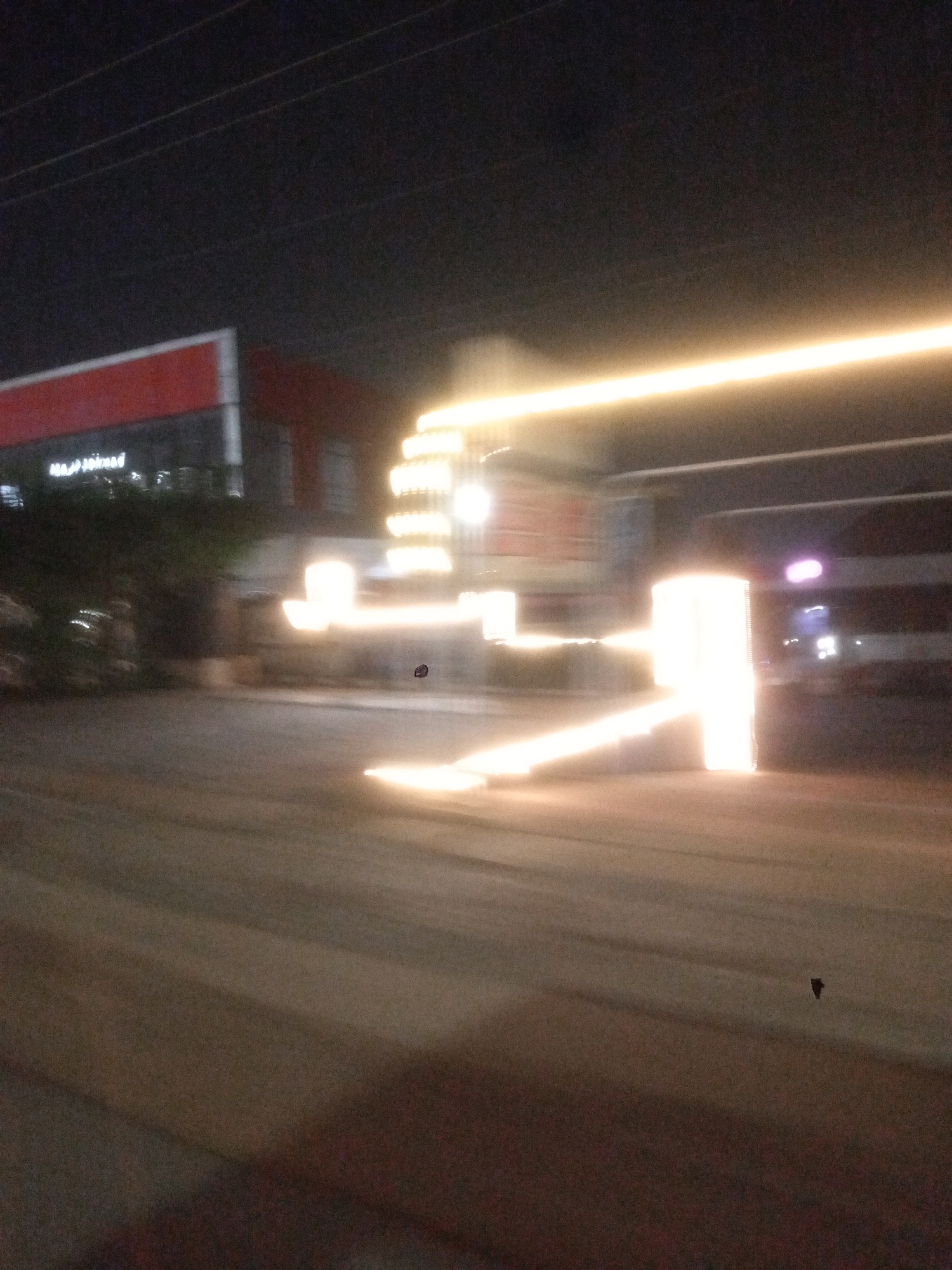 The image appears to be a nighttime long-exposure photograph taken in Gwarinpa, Nigeria. A blurred building with a red and white exterior is visible on the left side. Further away, a structure with numerous lights is apparent, resulting in streaks of light across the image. The foreground shows a blurred street surface. There are also power lines visible in the upper portion of the image.