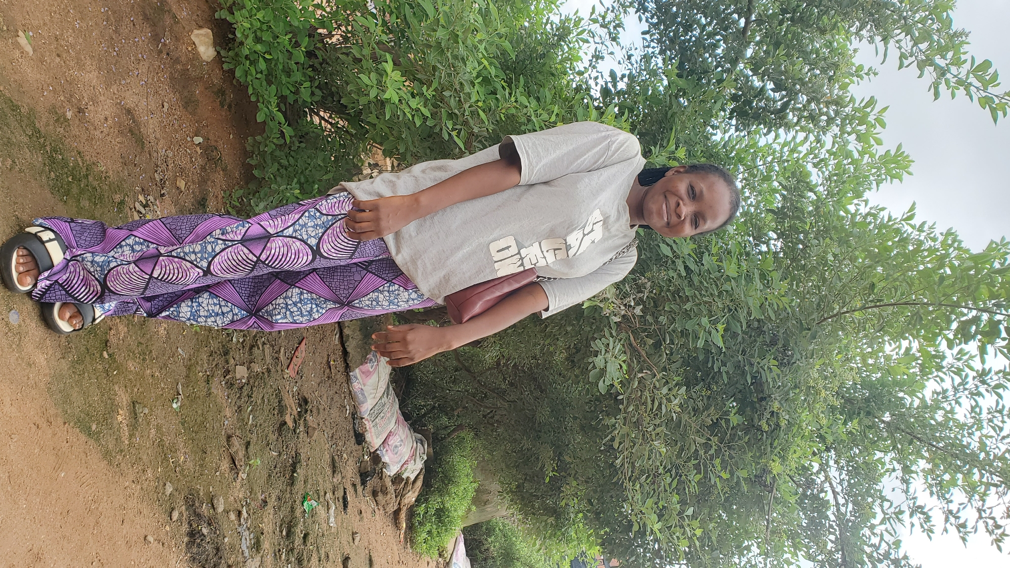 A young Black woman stands in front of a large green tree in a rural or semi-urban setting in Jalingo, Nigeria. She is wearing a light gray t-shirt with white lettering and loose-fitting purple pants with a geometric pattern. She has a maroon purse slung over her right shoulder. The ground is dirt and pebbles with some vegetation and a few bagged items, possibly construction materials, scattered in the background. The sky is overcast. The woman is smiling at the camera.