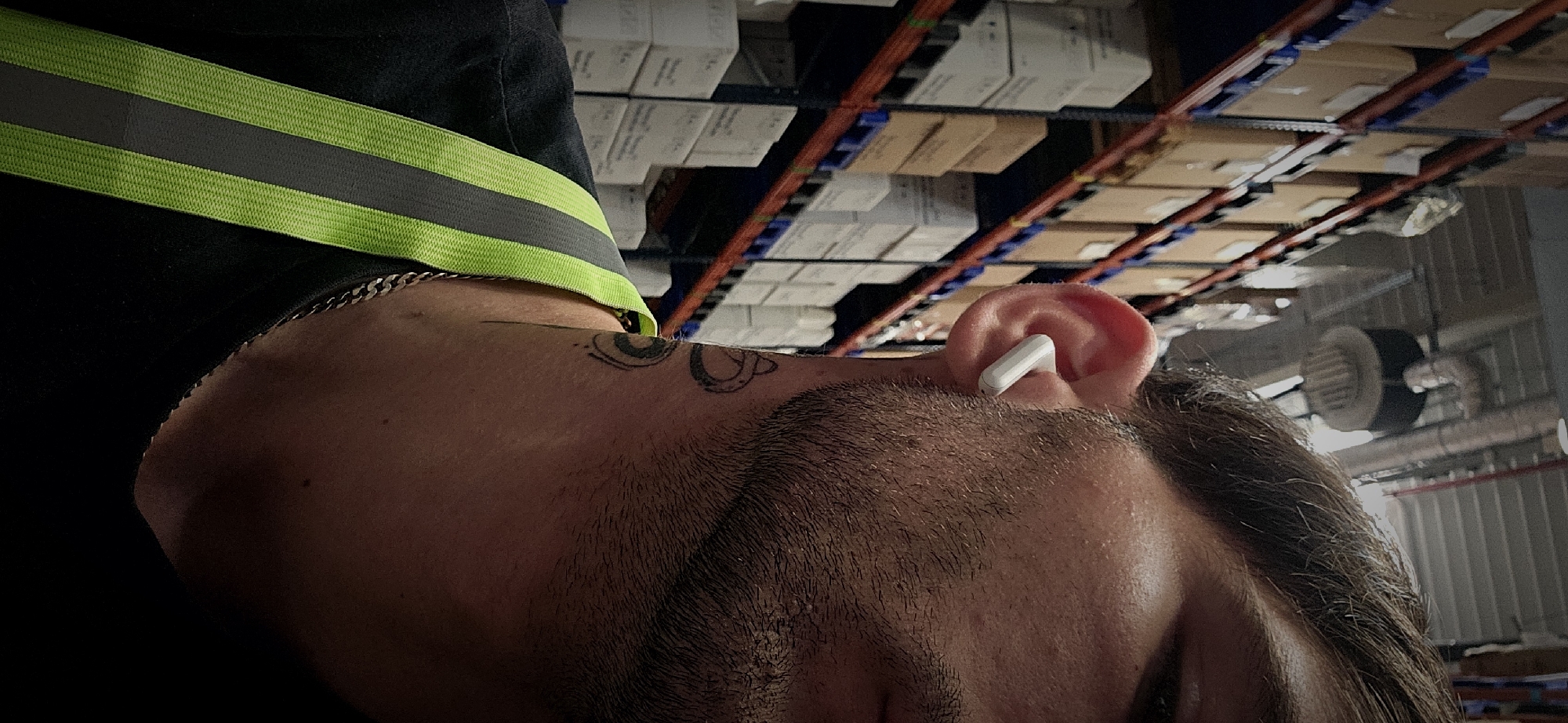 The image shows a man wearing a high-visibility vest with a lime green and grey stripe over a black shirt. He has a silver chain around his neck and tattoos near his hairline above his right ear. A white wireless earbud is in his ear. The background features warehouse shelving with multiple rows of boxes. It is not possible to determine if this is in Plovdiv, Bulgaria.