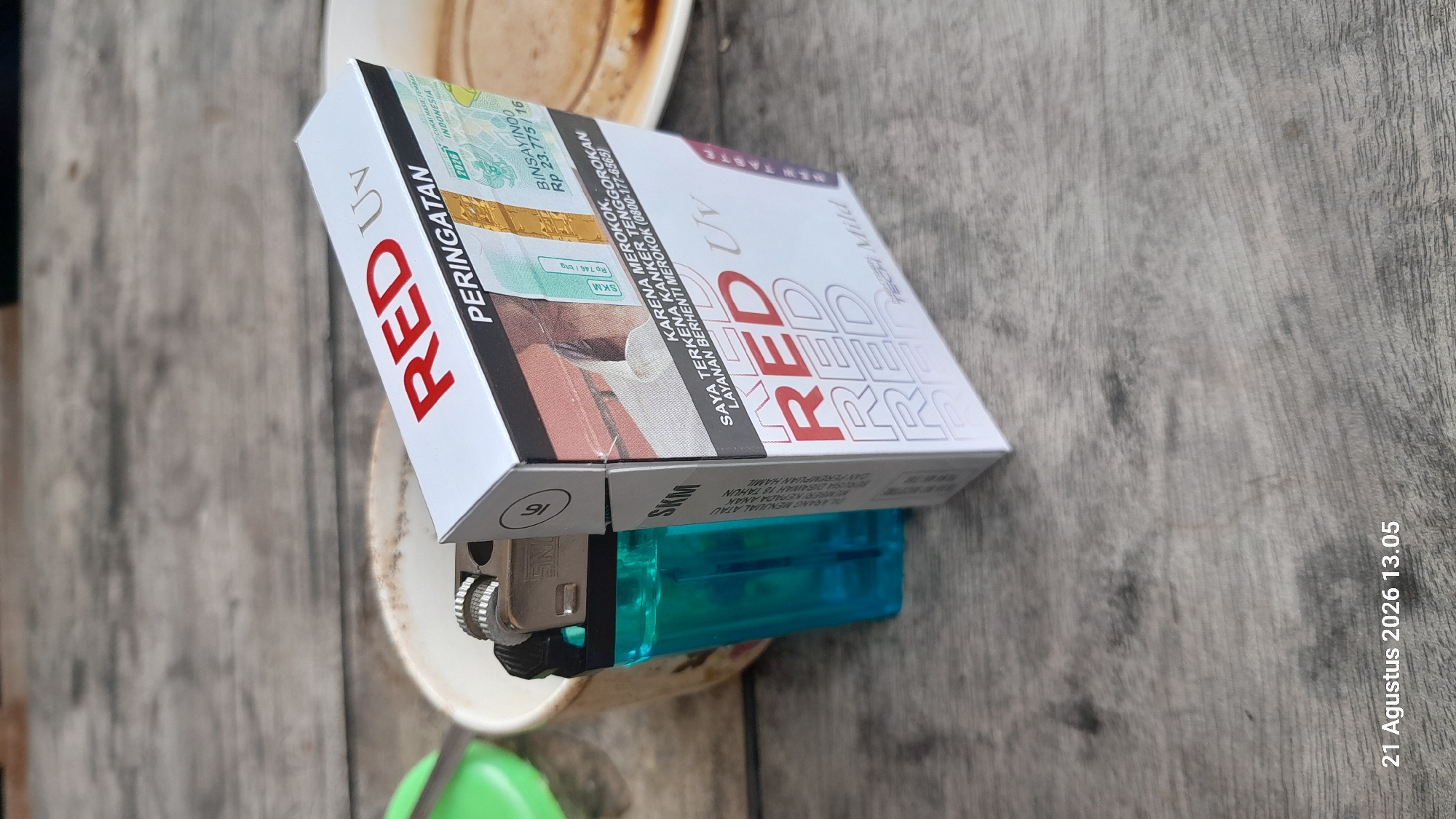 A cigarette pack of "RED UV" is displayed on a wooden surface, alongside a blue lighter and a white mug. The cigarette pack has the word "PERINGATAN" (Warning) printed on it, along with health warnings and tax stamps. The background shows a wooden table, suggesting an indoor or outdoor setting. The image is dated "21 Agustus 2026 13.05", which translates to August 21, 2026, at 1:05 PM. There is no discernible information about Takeran, Indonesia.