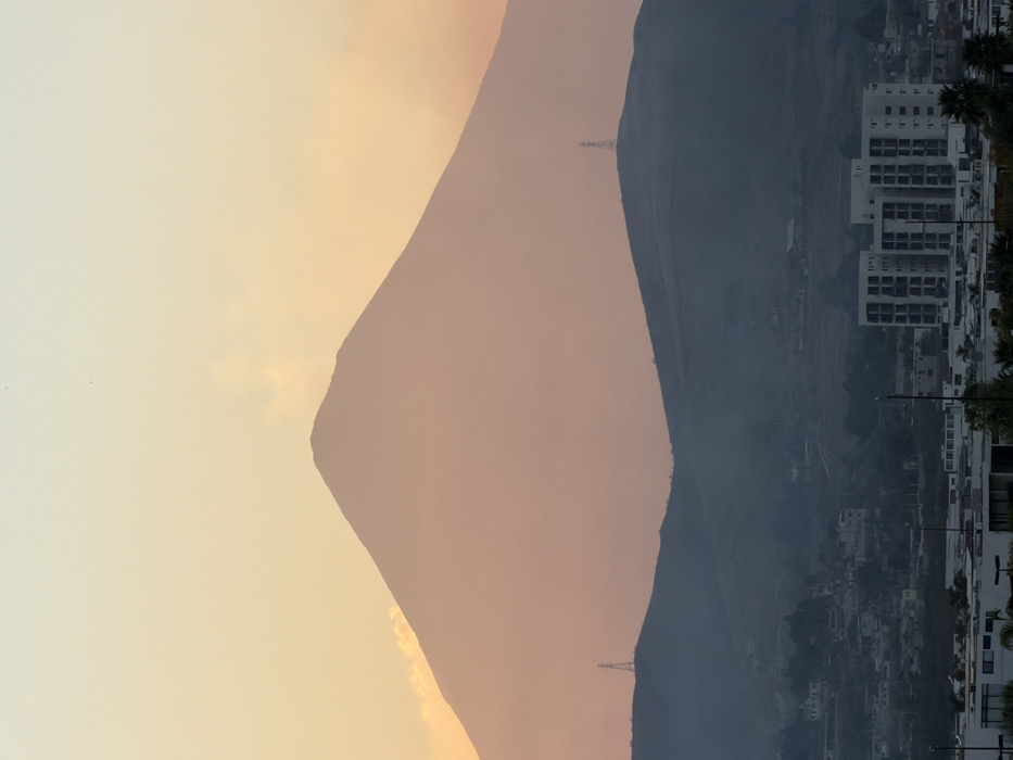 The image shows mountains at sunset or sunrise. The left mountain is pale orange, while the right mountain is dark gray. Power lines are visible on both mountains. A city stretches along the base of the right mountain. Buildings are white, and palm trees line the streets. The sky in the background is a soft orange hue.