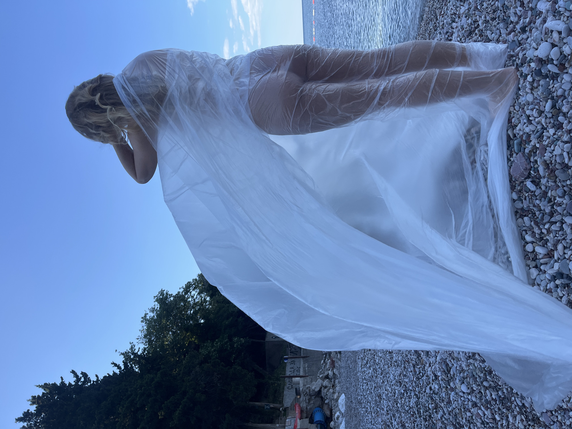 A woman lies supine on a pebble beach at Boreti, Montenegro, encased in a clear plastic material. The individual's body is visible through the plastic, and the head is turned with arms placed near the face. The backdrop is a coastal scene with a combination of trees, sky, and sea. The image is rotated.