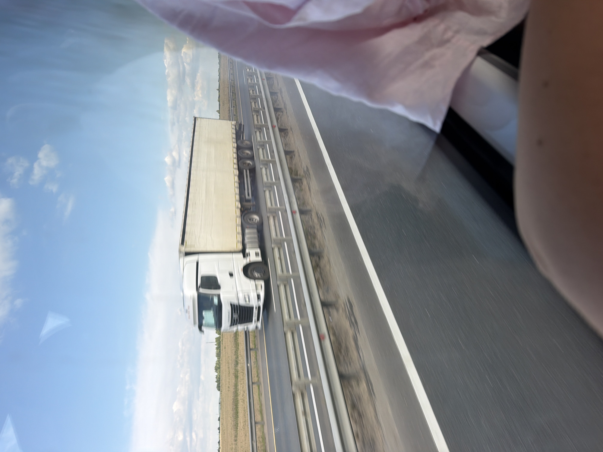 This image depicts a white semi-trailer truck driving on a highway. The truck is in motion, indicated by motion blur. The truck is white with a light-colored trailer. It is traveling on a multi-lane highway with a guardrail separating the lanes. The highway appears to be in a rural or semi-rural setting, with fields visible in the background. The sky is blue with scattered clouds. A portion of a pink garment and a person's arm are visible in the foreground, suggesting the photo was taken from inside a vehicle. The location is stated to be Pyatnitsa, Russia, though this cannot be independently verified from the image alone.