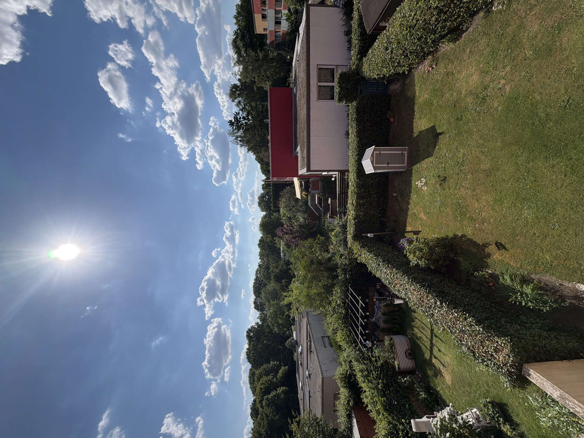 The image depicts a backyard garden scene in Dorsten, Germany. The viewpoint is from above, looking down and across a residential area. The primary subjects are the garden, a small shed, and a portion of a house.
The garden is well-maintained, with a lush green lawn and a thick, neatly trimmed hedge forming a boundary. A small, modern-looking shed with a light-colored roof and dark doors is positioned on the lawn, casting a long shadow, indicating it is likely afternoon. The background features dense trees and some residential buildings, including a building with a red facade.
The sky is a bright blue with scattered white, puffy clouds. The sun is visible in the upper left portion of the image, shining brightly and creating lens flare. This suggests it is a sunny day, likely midday or early afternoon.
The scene appears peaceful and tranquil, capturing a typical summer day in a suburban or semi-rural environment. There is no visible text in the image.