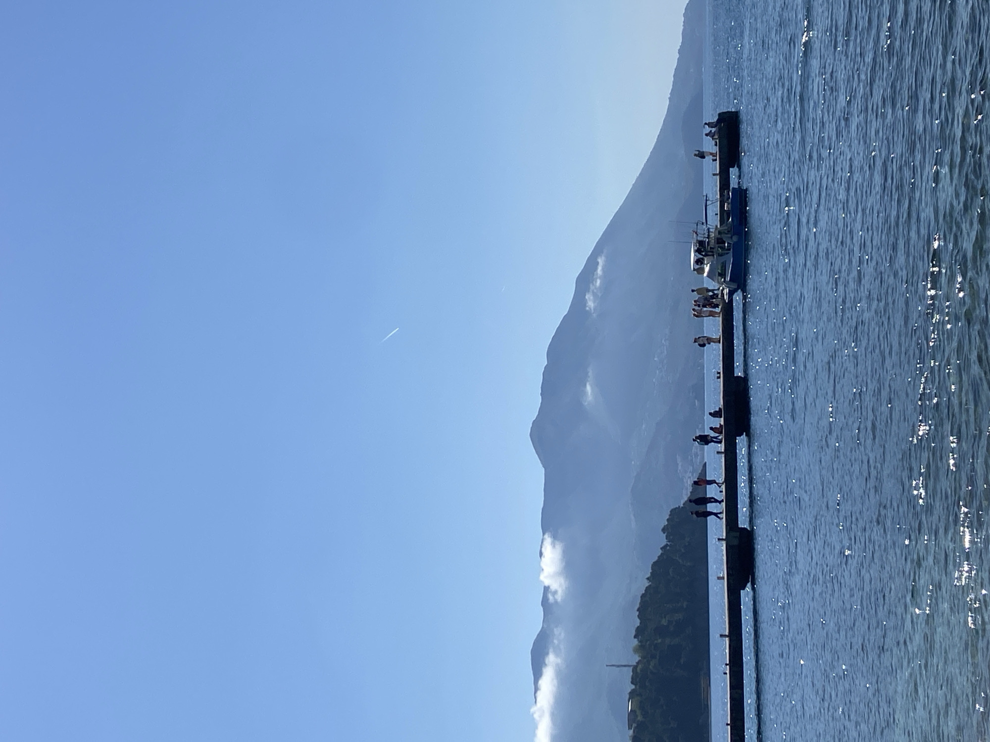 The image depicts a waterfront scene in Budva, Montenegro, with a long pier extending into the blue water. Silhouetted figures stand and sit along the pier. A small boat is moored on the pier. In the background, a mountain range rises, partially obscured by clouds. The sky is clear and blue. A thin contrail stretches across the sky.