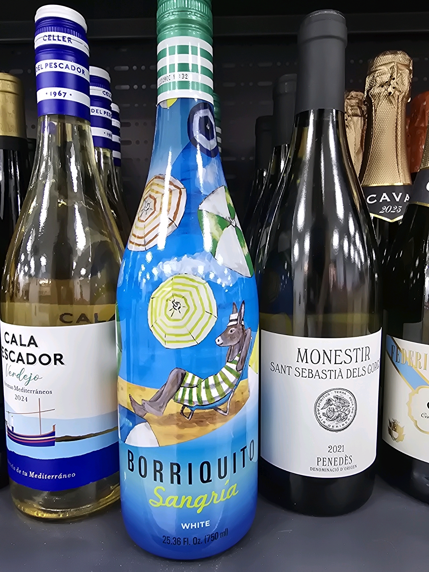 The image displays a shelf of wine bottles in what appears to be a retail setting, possibly a supermarket or wine shop. The bottles are arranged in a row, with the focus on several distinct labels.
On the left, a bottle labeled "CALA ESCADOR Verdejo" is visible. It's a clear glass bottle containing a pale yellow liquid. The label has white and blue elements, and a depiction of a sailboat. Above it, another bottle with a blue and white striped cap and "CELLER DEL PESCADOR 1967" is partially visible.
To the right of the Cala Escador bottle is a vibrantly decorated bottle of "BORRIQUITO Sangria WHITE." This bottle features a colorful illustration of a donkey relaxing on a beach under umbrellas, set against a bright blue sky. The text "BORRIQUITO Sangria" is prominently displayed in large letters, with "WHITE" in smaller print below.
Further to the right, a dark glass bottle with a black capsule is labeled "MONESTIR SANT SEBASTIÀ DELS GORGS PENEDÈS 2021." It has a white label with a circular emblem. Next to it, another bottle with a gold foil cap, identified as "CAVA 2023," is partially visible.
The background consists of dark shelving, common in retail environments. The lighting suggests an indoor setting with overhead illumination, creating reflections on the glass bottles.
There are no people or animals present in the image, and no actions or interactions are depicted. The environment is static and focused on the product display.
This image does not provide any information that would allow for its location to be identified within the city of Jaén, Spain.