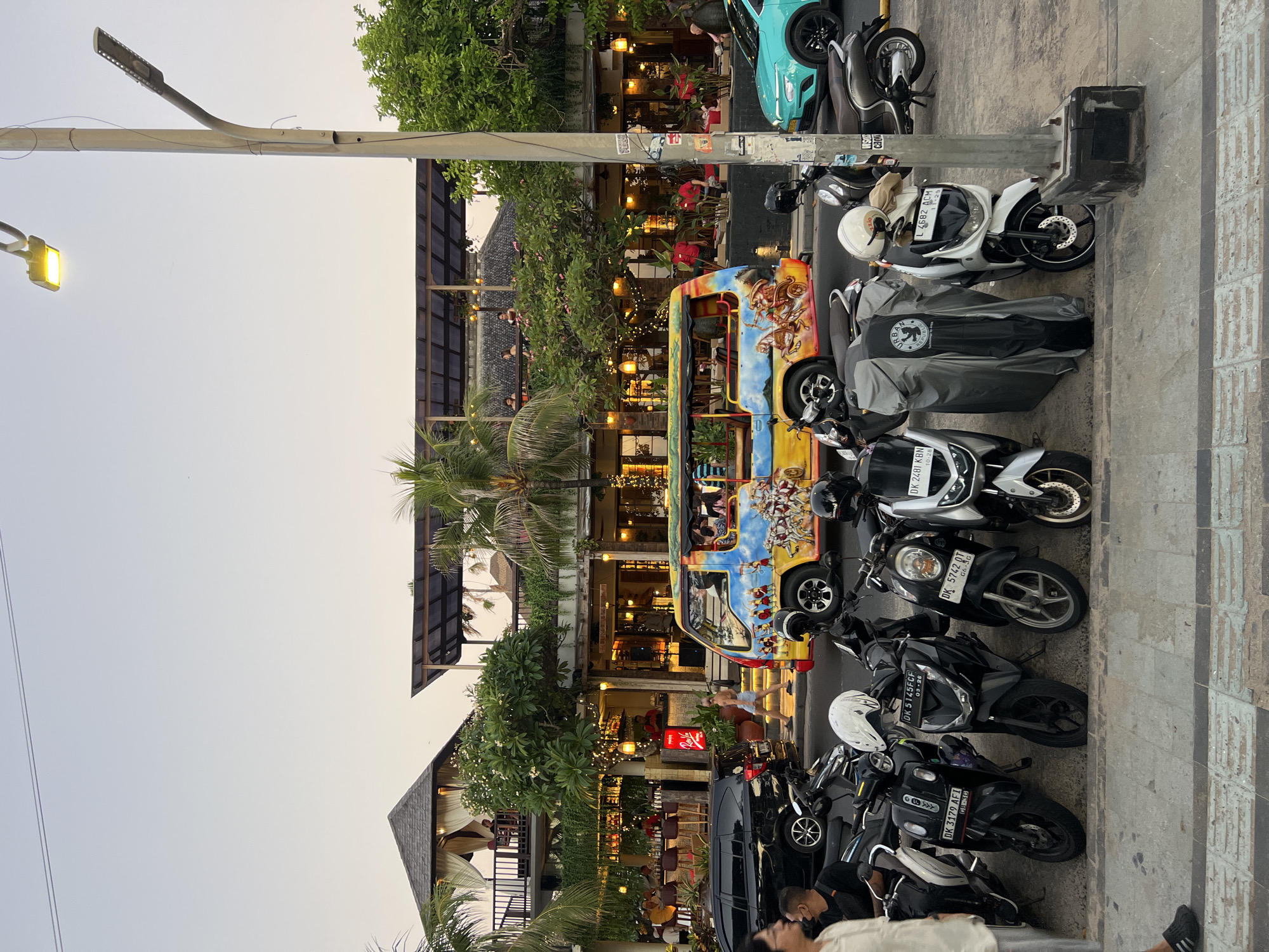 The image depicts a street scene in Kuta, Indonesia, likely during the late afternoon or early evening, given the warm light and streetlights beginning to illuminate. In the foreground, a row of parked scooters and motorcycles lines the paved sidewalk. Several of these vehicles are covered with protective grey tarps. To the right, a teal-colored car is parked.
The middle ground features a colorful, open-air van, decorated with depictions of people and horses, driving down the street. Behind the van, a two-story building with balconies and open-air dining areas is visible, decorated with strings of warm lights. People are seated at tables on the upper level and inside the building. Several palm trees are interspersed amongst the structures, and lush greenery is present. A black SUV is parked on the left side of the street.
In the immediate foreground, on the left side of the image, a person wearing glasses and a light-colored shirt is partially visible, walking away from the camera. Another person is visible behind them, wearing a dark t-shirt. The environment appears to be a commercial area, possibly a street lined with restaurants or shops. A tall lamppost stands prominently on the right side of the frame, with one of its lights illuminated. The sky is a pale orange, suggesting dusk.