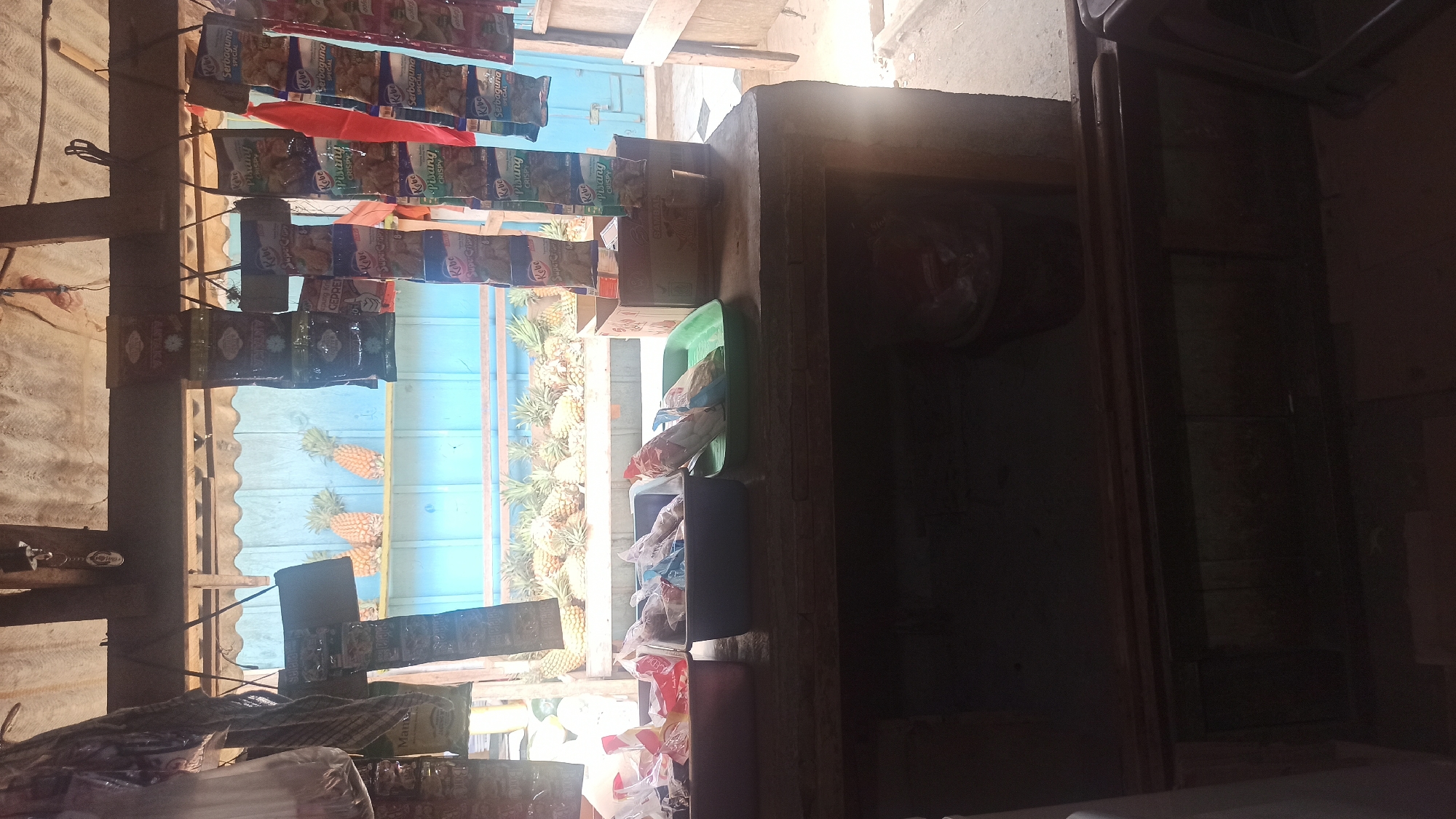 This is a daylight, medium-shot, eye-level photograph of a small roadside stall in Tuba, Indonesia. The stall is displaying various items for sale, including packaged snacks, and a large quantity of pineapples. The stall appears to be constructed from wood and metal, with a corrugated metal roof. The stall is open to the elements, with a blue wall visible in the background. The foreground shows a wooden counter with three plastic containers holding packaged goods. Behind the counter, a shelf displays numerous pineapples. Packaged snacks hang from the ceiling of the stall. The scene is brightly lit, with strong sunlight casting shadows.