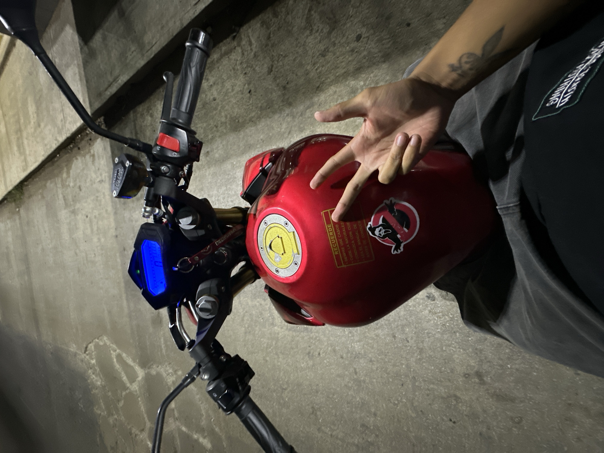 The image displays an overhead perspective of a person seated on a red motorcycle at night in Macho Muerto, Venezuela. The motorcycle's red fuel tank is prominent, featuring a yellow fuel cap with six visible fasteners. A rectangular sticker on the tank contains text in Spanish, partially legible, including "RECUERDE" (Remember), "SIEMPRE USE CASCO" (Always use a helmet), "CUIDE LA NATURALEZA" (Care for nature), "CONDUZCA CON CUIDADO" (Drive carefully), and "LEA EL MANUAL" (Read the manual). Below this is a circular sticker depicting the "Ghostface" character from the *Scream* franchise within a red circle with a diagonal line, mimicking the Ghostbusters logo.
The handlebars are visible, with a blue-illuminated digital dashboard displaying "0.0" and a green indicator light. A key with a red lanyard is inserted near the ignition. The right handlebar includes the grip, brake lever, and a translucent brake fluid reservoir. The left handlebar shows the grip and control buttons.
In the lower right of the frame, the person's right arm and hand are visible. The hand is positioned with the thumb, index, and middle fingers extended, while the ring and pinky fingers are folded. The individual wears a dark-colored short-sleeved shirt with a partially visible white and black graphic on the chest and dark pants. A tattoo resembling a stylized winged figure is visible on the inner right forearm. The ground surface is paved, exhibiting texture and some cracks, illuminated by artificial light sources.