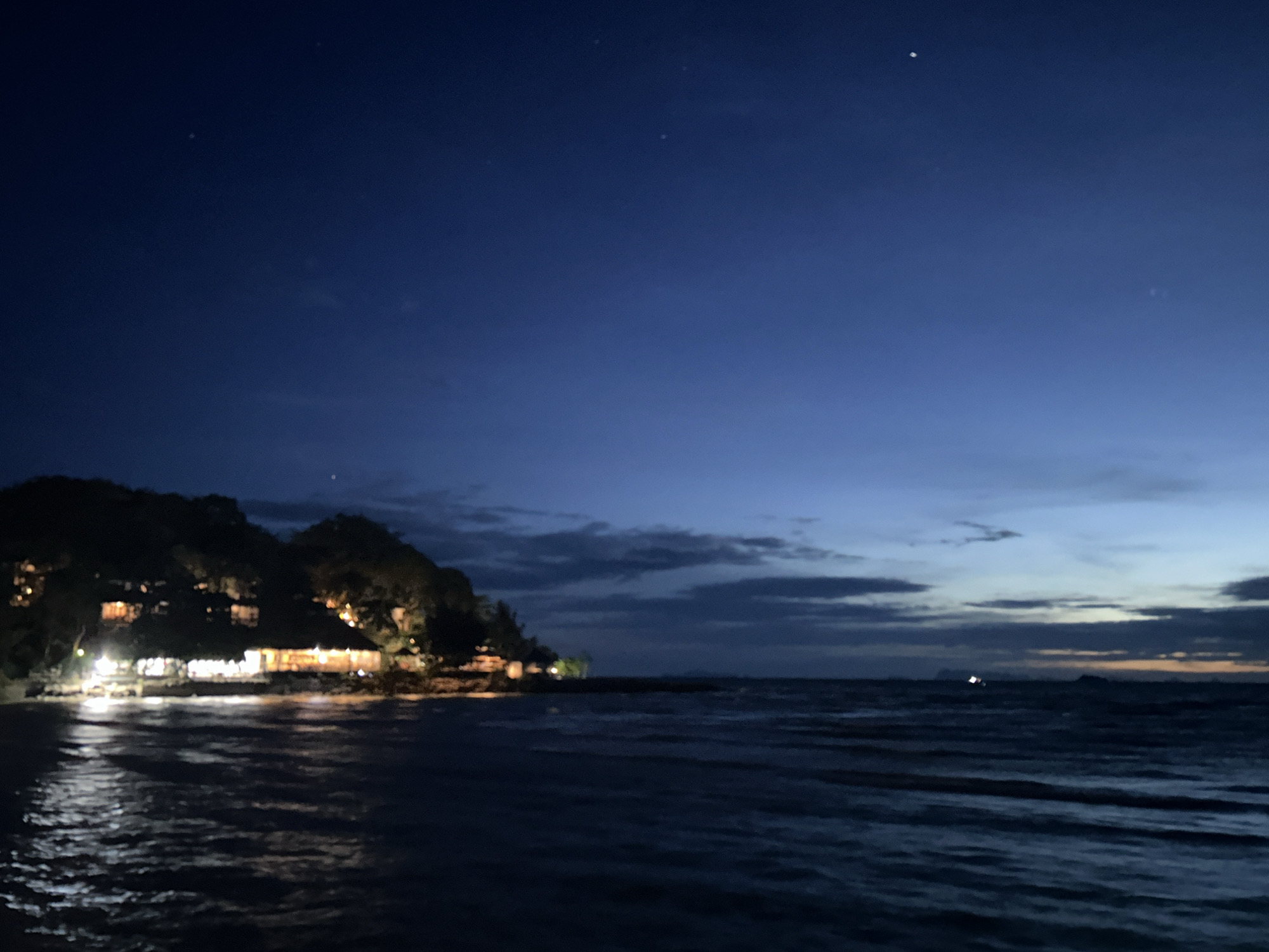 Night view of a beach at Baan Sri Thanu, Thailand. A row of illuminated buildings is situated along the beach at the base of a forested hill. The dark water is lightly illuminated by reflections from the lights. The sky transitions from dark blue above to slightly lighter shades near the horizon, with some low clouds present. A few stars are visible in the upper portion of the sky.