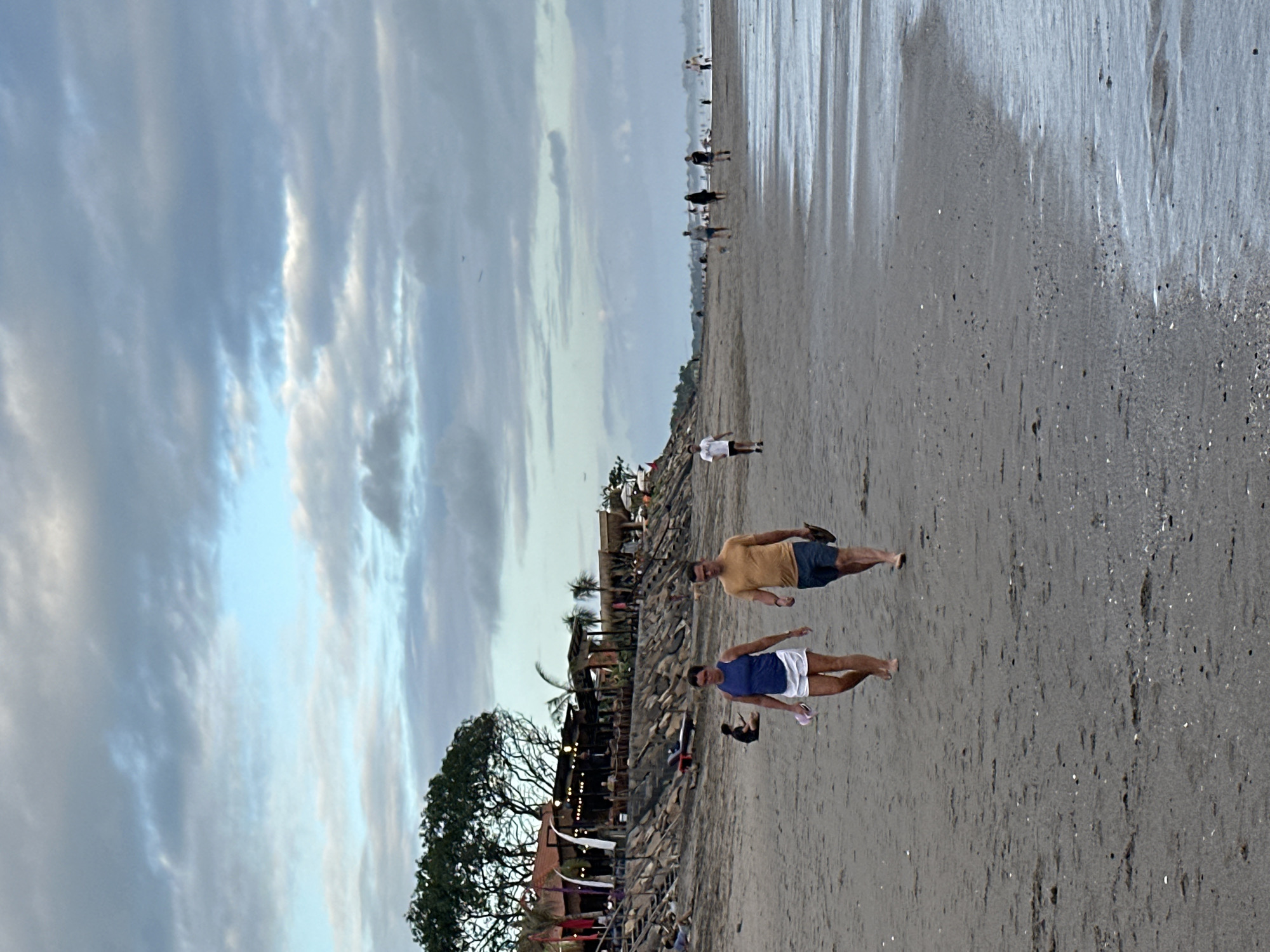 In the city of North Kuta, Indonesia, a beach scene unfolds under a cloudy sky. The tide is low, leaving wet sand that reflects the overcast sky. Several people are scattered along the beach. In the foreground, a couple walks towards the camera. The woman wears a blue tank top and white shorts, carrying a pair of flip-flops. The man beside her is in a yellow t-shirt and blue shorts, also carrying flip-flops. Further down the beach, other individuals are visible, some walking, others sitting or standing. A restaurant structure with thatched roofs and lights is situated on a rocky embankment behind the beach, lined with palm trees and foliage. The overall atmosphere suggests a tranquil evening on the coast.
