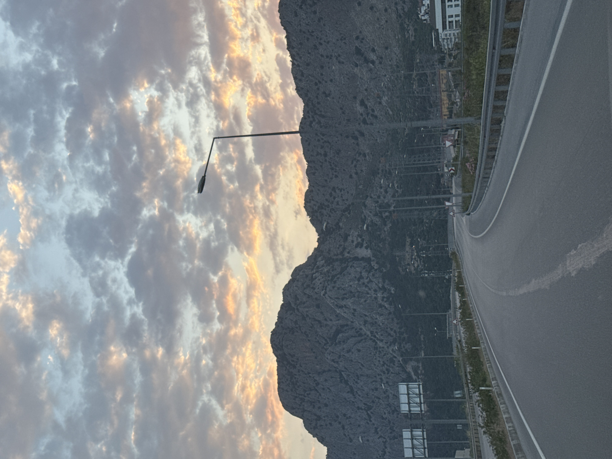 This image depicts a scenic landscape featuring a multi-lane road winding alongside a rugged mountain range, set against a dynamic sky, likely captured in Konyaaltı, Turkey. The time of day appears to be either sunrise or sunset, indicated by the warm, golden and orange light illuminating patches within the otherwise grey and blue clouds that fill the left side of the frame.
The central subject is a dark, rocky mountain, characterized by its uneven and imposing silhouette, which dominates the mid-ground. A tall, single streetlight with a curved arm stands prominently against the cloudy sky and the mountain's edge. To the right, a smooth asphalt road with a clear white lane divider curves gently. A metallic guardrail separates the road from the lower slopes of the mountain, where some sparse greenery is visible.
Along the road and extending up the mountain's side, various utility poles or infrastructure elements can be seen. In the upper right corner, beyond the road, faint outlines of buildings or structures suggest a distant urban presence. The scene is devoid of people or vehicles, conveying a tranquil and expansive atmosphere. There is no legible text visible within the image.