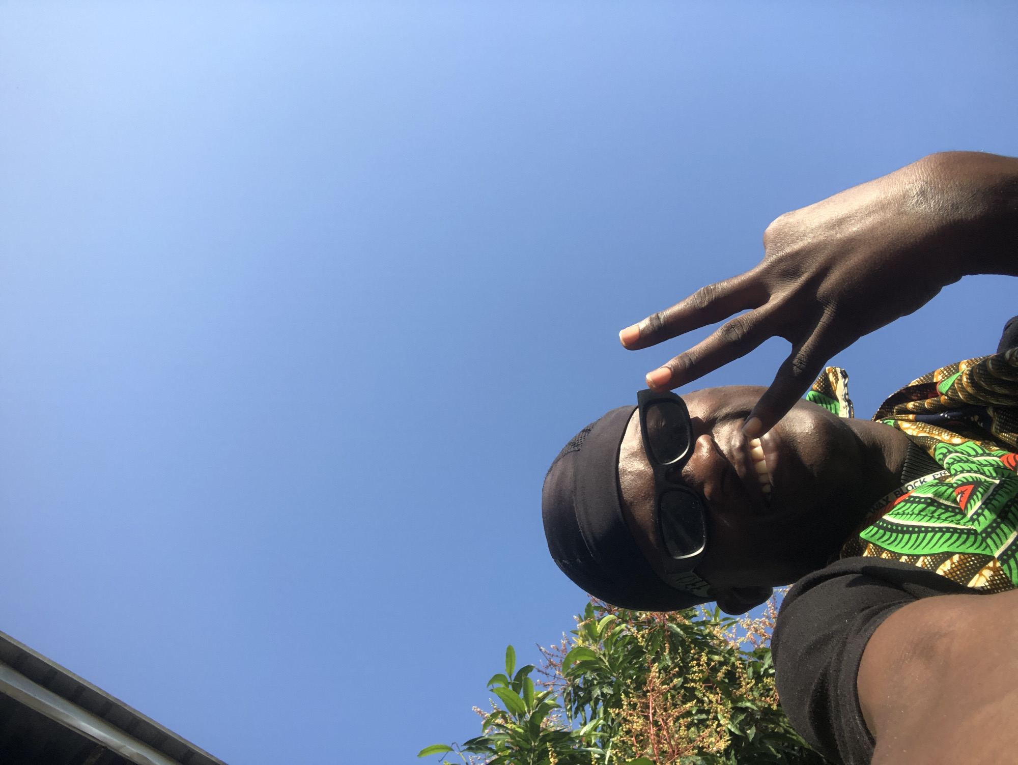 In Ado, Nigeria, under a clear blue sky, a person wearing a black durag and black sunglasses is captured in a low-angle selfie. They are dressed in a black t-shirt and a vibrantly patterned green, yellow, and black shirt or scarf around their neck. The individual smiles broadly, showing their teeth, and holds up two fingers in a peace sign. To the left of the frame, lush green foliage from a tree is visible against the bright sky, and a portion of an overhead structure, possibly a roof or overhang, is visible in the upper left corner.