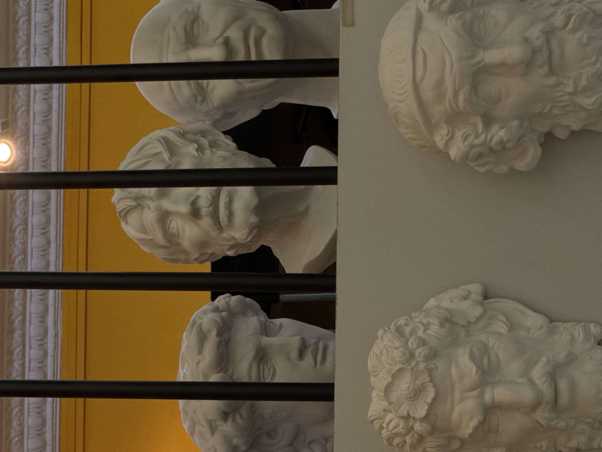 The image depicts a display of white plaster busts of various male figures, arranged in two tiers. The upper tier features three busts positioned behind dark vertical bars, with a yellow wall and ornate molding visible in the background. The busts appear to be of classical or historical figures, characterized by their detailed sculpted features, including hair, beards, and facial expressions.
The lower tier displays two more busts, also made of white plaster, mounted on a plain, light-colored surface. These busts also exhibit intricate sculpting of hair, beards, and facial features, with one adorned with a floral headdress.
The scene is indoors, likely in a museum or gallery setting, judging by the presentation of the busts as exhibits. There are no people visible in the image. The lighting appears to be artificial, illuminating the sculptures. The overall impression is one of an artistic or historical exhibition. The location of this display in Moscow, Russia is not identifiable from the visual information provided in the image.