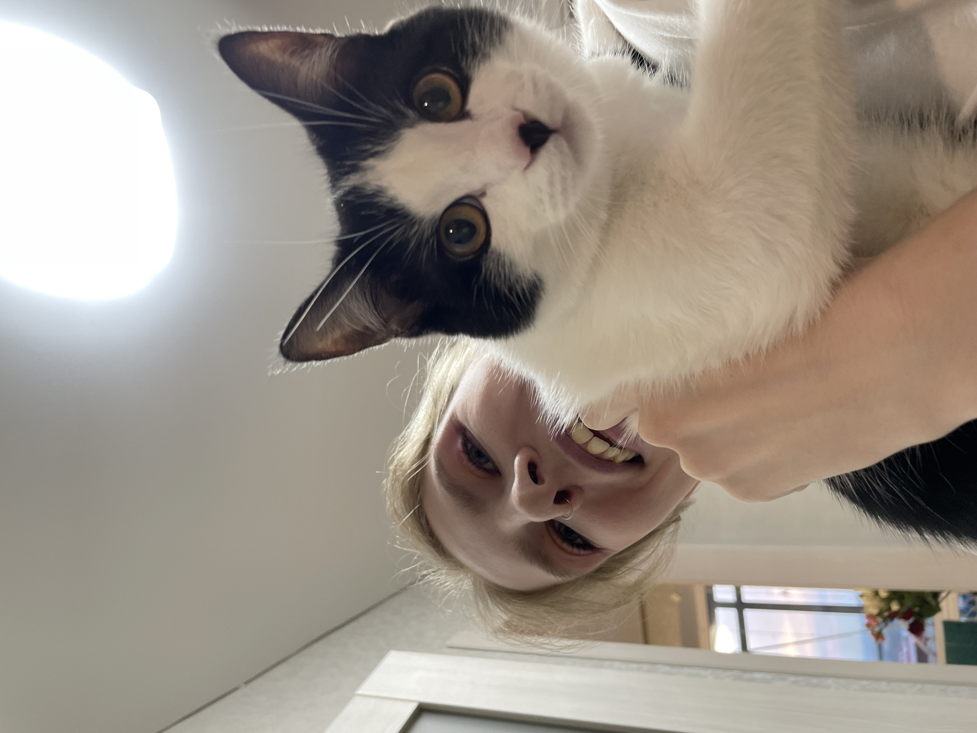 The image is taken from a low-angle, upward-facing perspective. A person with fair skin and blonde hair holds a black and white cat. The cat has yellow eyes and a black nose. The person is smiling, revealing their teeth. Behind the person and cat, a partially visible window shows the sky and a building with decorations. The location appears to be indoors, and the background suggests an interior space, potentially a room with a door and a white ceiling. There is no explicit information in the image to confirm that the location is Izhevsk, Russia.