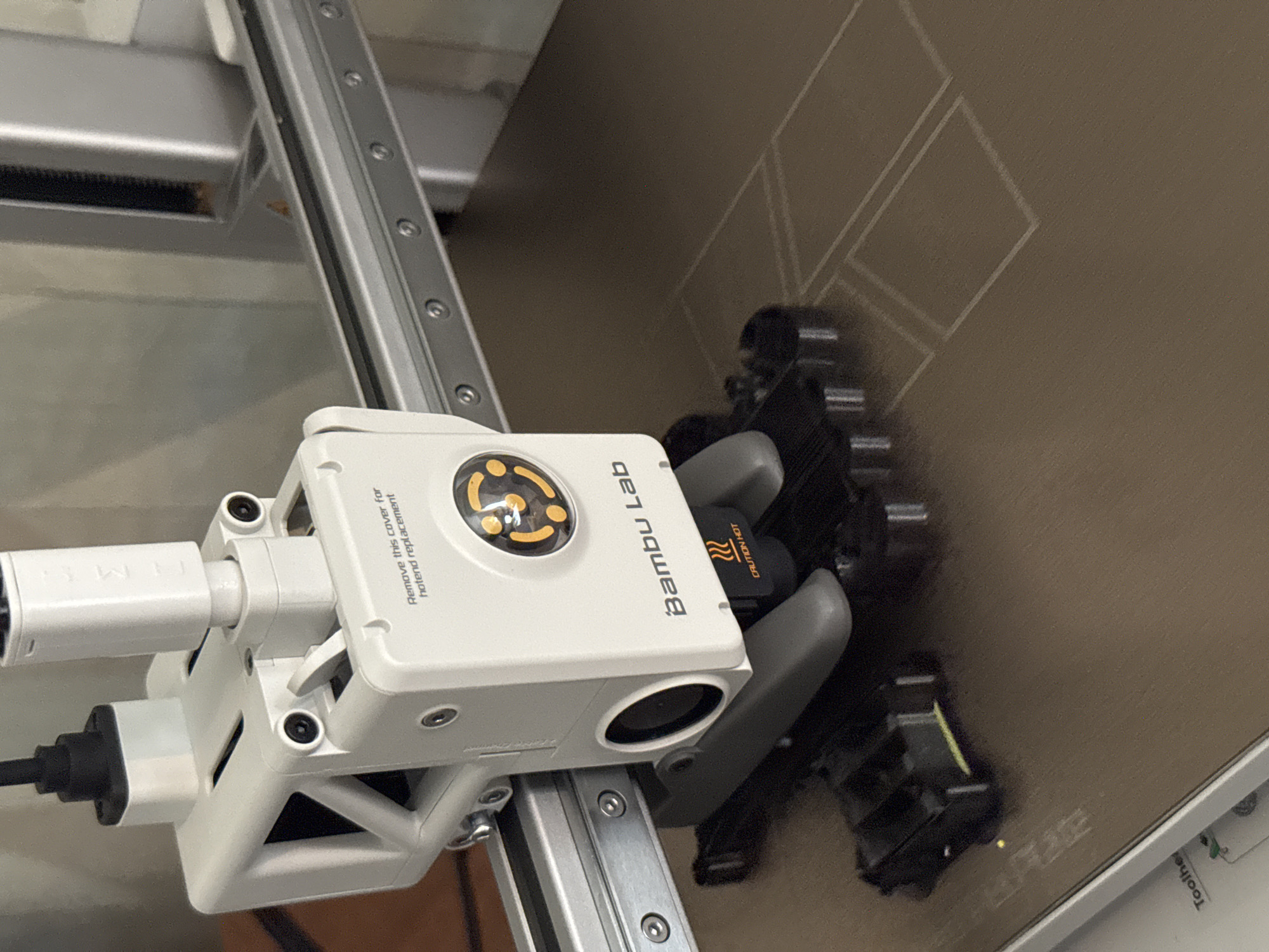 The image shows a close-up of a white 3D printer head, labeled "Bambu Lab," positioned above a textured brown print bed. The print bed has several thin, white lines etched onto it, forming geometric shapes. Attached to the print head is a black component with yellow warning text that reads "CAUTION HOT." The printer is mounted on a silver metal rail system. The background is softly blurred, showing what appears to be a wooden surface and a larger silver structure. There are no people or animals visible in the image. The image does not contain any information that could be used to determine its location in the city of Белоус, Russia.