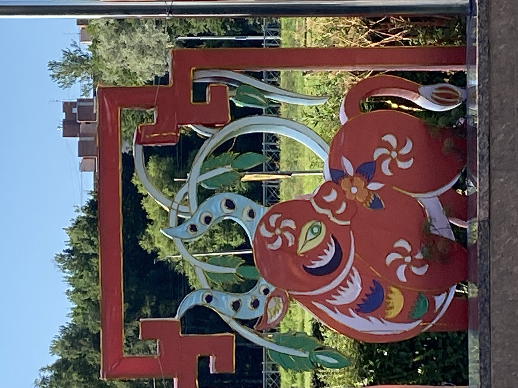 The image depicts a festive, stylized red ox sculpture as part of an outdoor decoration. The ox is adorned with white, blue, and yellow patterns, including floral designs and swirling motifs. Its horns are light blue with black dots. The sculpture is framed by a red, ornate structure with gold trim. Behind the sculpture, trees and buildings are visible under a clear blue sky. This artwork is located at the Nevsky Prospect shopping mall in Saint Petersburg, Russia.