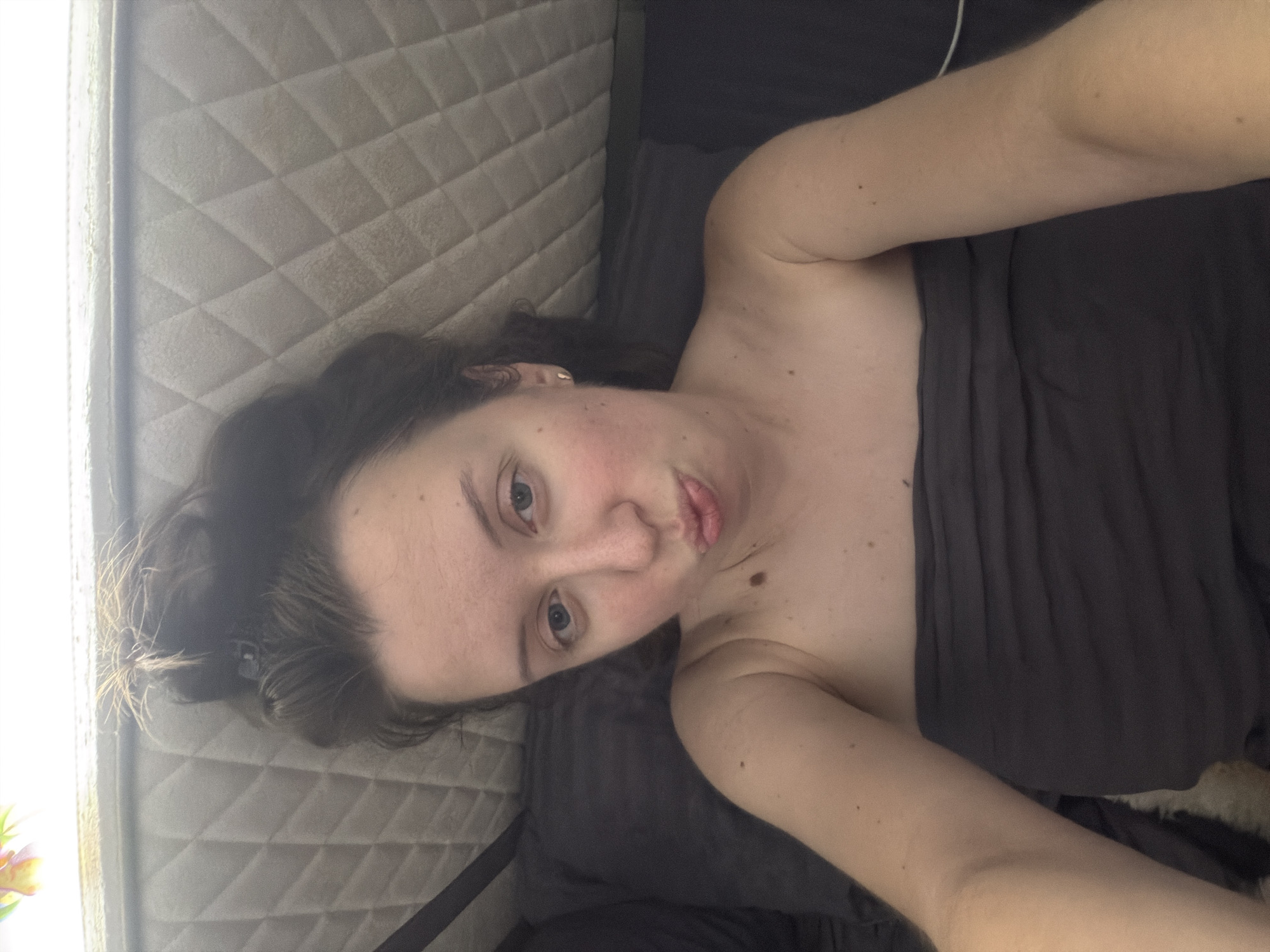A close-up selfie shows a young Caucasian woman lying in bed, with her head propped against a quilted headboard. She is wearing a dark grey strapless top and her hair is pulled up in a messy bun. The woman is looking at the camera with a playful expression, puckering her lips. Her skin is fair with some freckles and a mole on her left cheek. The room appears to be dimly lit, with natural light coming from the top of the image. The background consists of a dark pillow and the quilted headboard of a bed. There are no visible indications of the location being in Bila Tserkva, Ukraine.