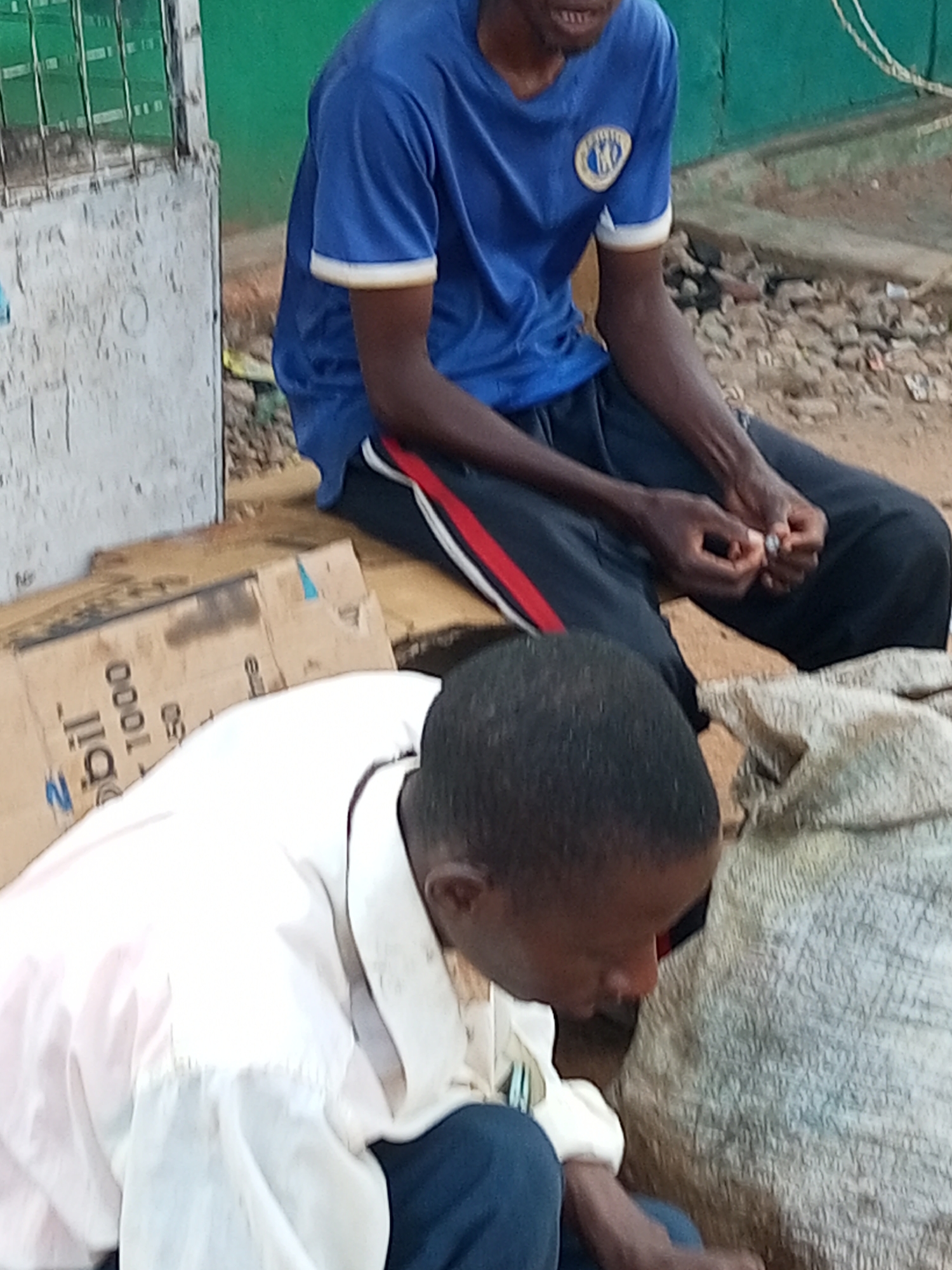 Two young men are sitting outdoors, possibly in Nigeria. The man in the foreground is wearing a white collared shirt and dark pants, with his head bowed as if concentrating on something in his lap. His skin tone is dark, and he has short, dark hair. The man in the background is wearing a blue t-shirt with a white and yellow emblem on the chest and dark pants with a red stripe down the side. He has his hands clasped in front of him, and is looking downwards.
The scene appears to be informal, with a rough, possibly dirt ground and some scattered rocks. There is a piece of cardboard in the foreground with some text on it. The visible text reads "obil 1000 50". The overall lighting suggests it is daytime. The setting appears to be a somewhat urban or semi-urban area, given the presence of what looks like a concrete structure with a green surface and a metal fence in the background, as well as the cardboard. The mood is neutral, with no strong indicators of specific emotions.