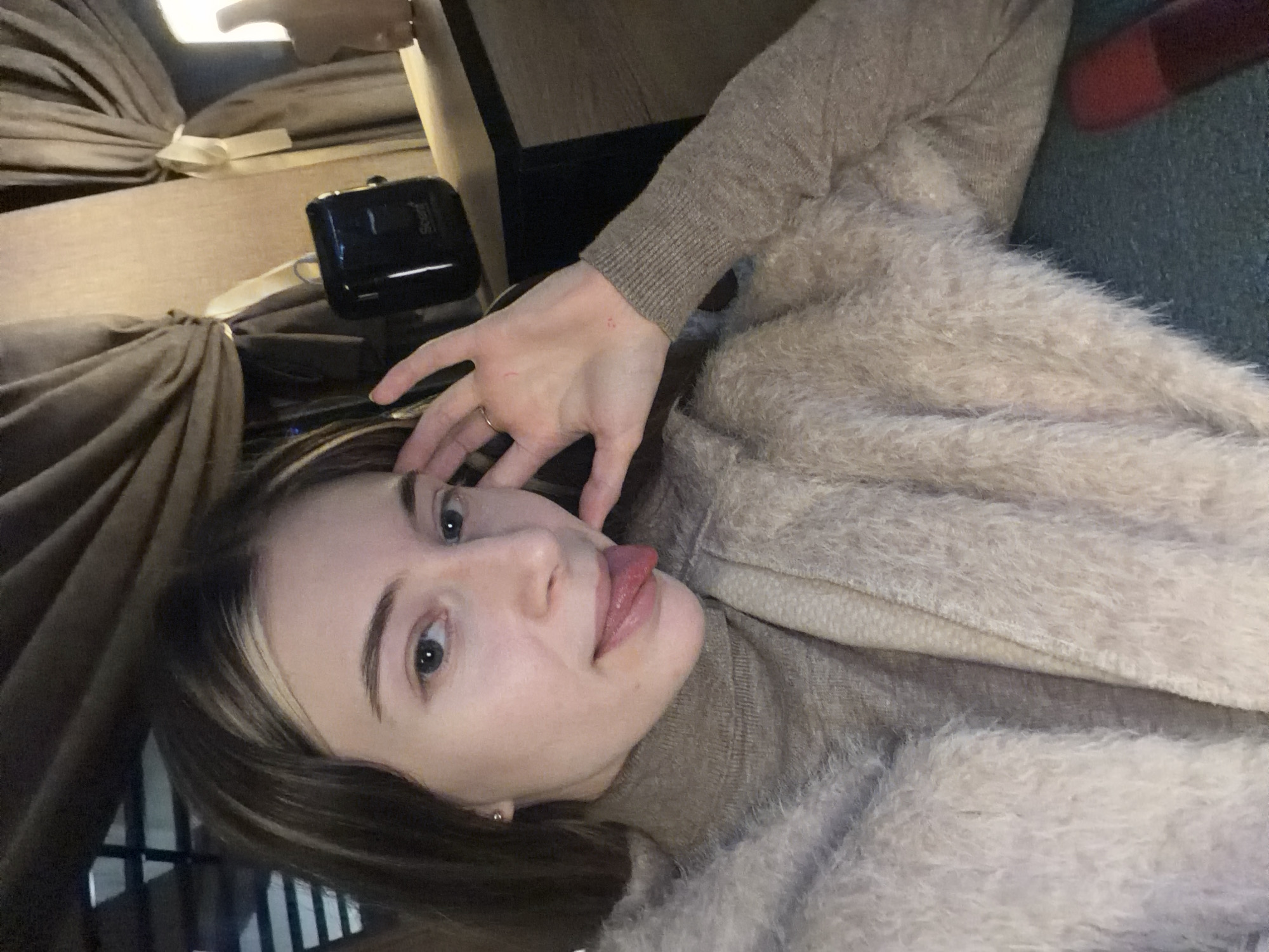 A young woman is lying down, possibly indoors, given the presence of a tan-colored curtain and light fixture in the background. She is wearing a light brown turtleneck sweater underneath a fuzzy, cream-colored jacket or sweater. Her hair is brown with blonde highlights. She is sticking out her tongue while holding her hand near her face, with what appears to be a gold ring on one of her fingers. A black object with a cord attached sits behind her. The background is blurred, hindering a precise description of the location. The image does not provide any information regarding the city of Plovdiv, Bulgaria.