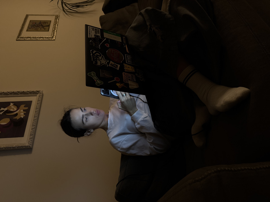 A woman is seated on a dark-colored sofa in a dimly lit residential interior within Venice, Italy. She has dark hair styled in a bun and wears a white long-sleeved shirt and dark pants. Her face is illuminated by the screen of a silver smartphone, which she holds with both hands. Her gaze is directed towards the phone. An open black laptop rests on her lap, its screen also emitting light. The laptop lid is adorned with various stickers, including one that reads "WE LOVE DOGS." Her lower legs are visible, covered by white socks with two blue stripes near the cuffs. In the background, a light-colored wall features two framed artworks: a larger, ornate white-framed piece depicting stylized, abstract figures in yellow and purple hues on the left, and a smaller, gold-framed artwork on the right. A green plant is partially visible behind the sofa on the far right. The primary light source in the scene emanates from the electronic device screens.