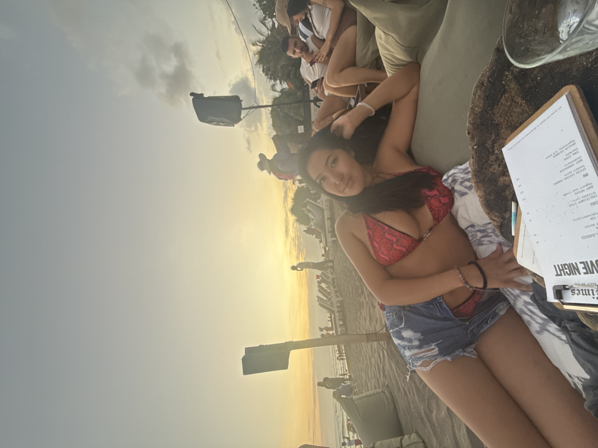 This image depicts a beach scene in North Kuta, Indonesia, during sunset. A young woman in a red bikini top and ripped denim shorts is lounging on a beanbag, looking towards the camera. Behind her, other beachgoers are visible, some sitting on lounge chairs, others walking along the shore. Two speakers are set up on wooden posts, and palm trees line the background. The sky is a gradient of orange and yellow, indicative of a sunset. In the foreground, a menu and a glass are placed on a wooden surface. The overall atmosphere suggests a relaxed evening at a beachside establishment.