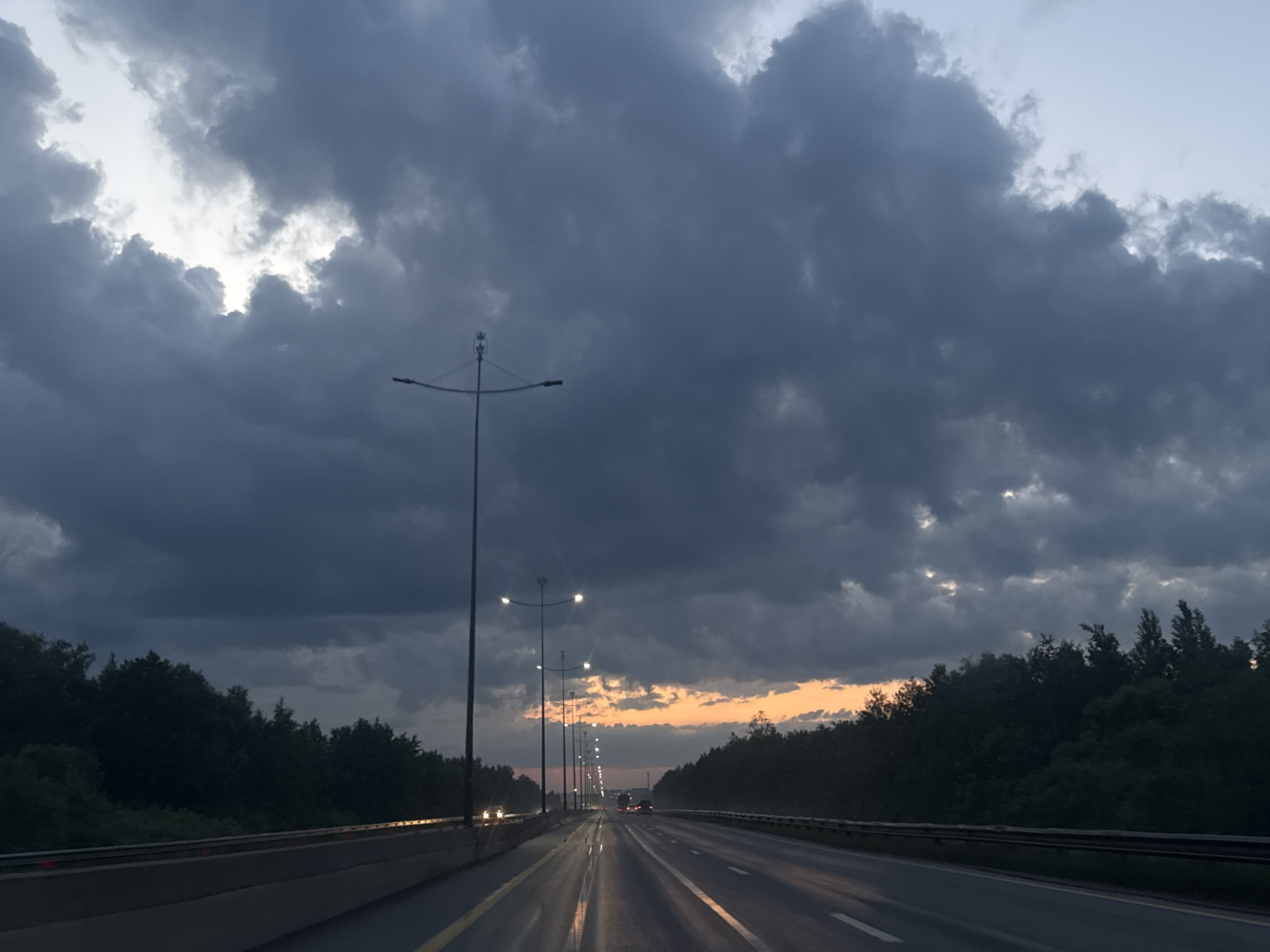 The image depicts a view from a vehicle traveling on a highway in the vicinity of Kondratovo, Russia, during a cloudy sunset. The sky is filled with dark, dramatic clouds, with a sliver of orange and yellow light visible near the horizon, indicating the sun setting. Trees line both sides of the highway, appearing dark against the twilight sky. Several streetlights are visible along the roadside, illuminating the wet asphalt of the highway. A few vehicles are seen in the distance, their headlights and taillights creating streaks of light on the road. The overall atmosphere is somber and atmospheric, suggestive of approaching dusk or a storm.