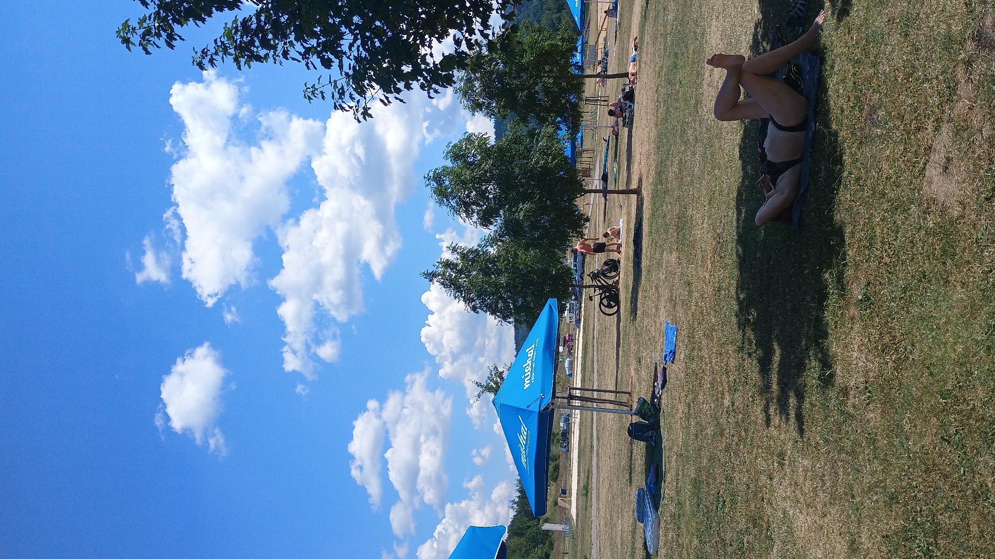 This is a daytime, eye-level, outdoor photograph of a grassy area in Sokolov, Czechia, likely a recreational park or beach area, under a partly cloudy blue sky. In the foreground, a person wearing a black bikini is lying on their back on a dark mat, resting in the shade of a tree. Their legs are bent and crossed at the ankles, and their arms are resting behind their head. To the left of the foreground, on the grass, are several items including a deflated air mattress, a backpack, and a blue towel. Further back, a large blue umbrella is set up, with the logo "michal" visible on it. Nearby, a man and a child are standing, and a bicycle with thick tires is parked next to them. In the mid-ground, several more trees are scattered across the grass, and in the background, a sandy area with some beach chairs and umbrellas is visible, along with parked vehicles and distant trees and hills. The overall scene depicts a leisurely summer day.