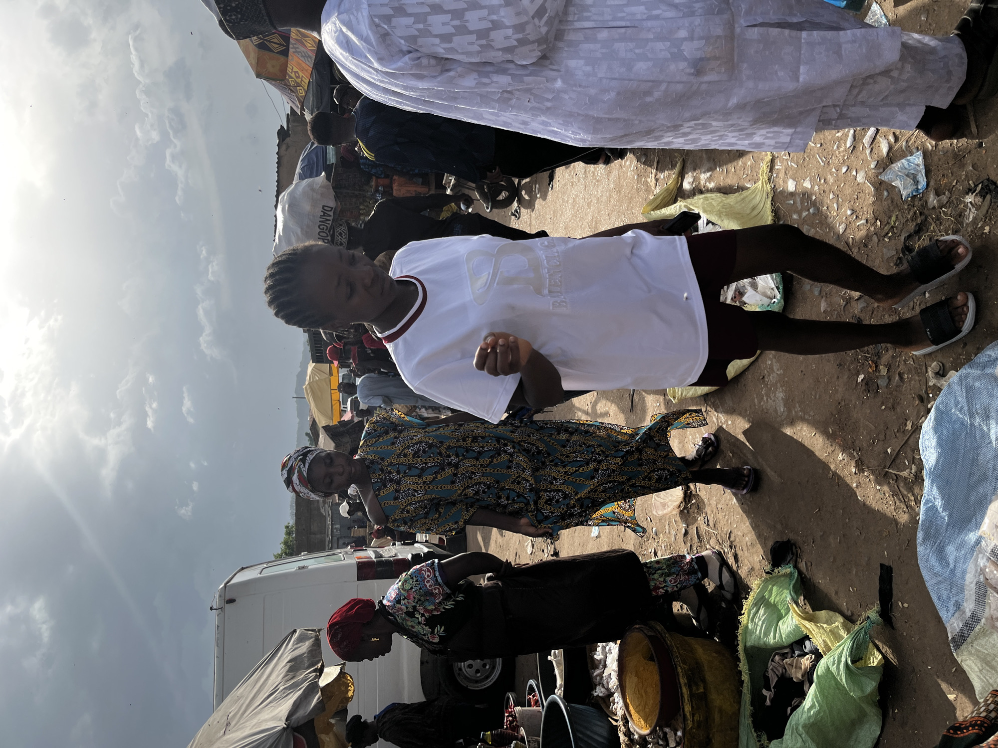 The image depicts an outdoor market scene in Rafin Par, Nigeria. A young woman in a white t-shirt and dark shorts walks forward, looking down. To her left, another woman in a colorful patterned dress and headscarf walks in the same direction. Behind them, a white van and various market stalls with awnings are visible. The ground is dirt and strewn with debris, including green and blue plastic bags and discarded items. Several other people are present in the background, some walking and others standing. The sky is overcast with dramatic clouds, and sunlight breaks through in places, casting shadows on the ground. The overall impression is one of a bustling, everyday market day.