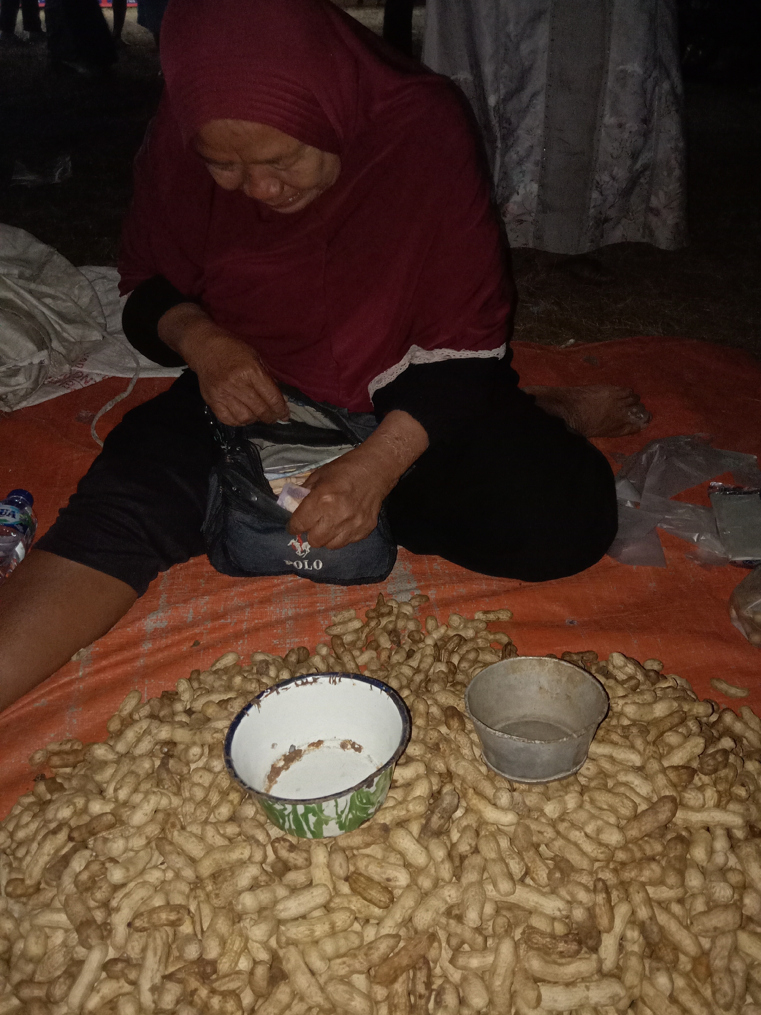 In Stiaji, Indonesia, a person wearing a maroon hijab and black pants is sitting on an orange tarp, sorting through a large pile of peanuts. The person is looking down at a dark blue pouch with "POLO" written on it, from which they are extracting what appears to be money. To the left of the person, a plastic water bottle with a blue label is visible. In the foreground, two bowls are placed among the peanuts; one is white with a green and blue rim, and the other is a small, grey metal bowl. The background is dark, with indistinct figures and a draped fabric, suggesting an outdoor setting at night.