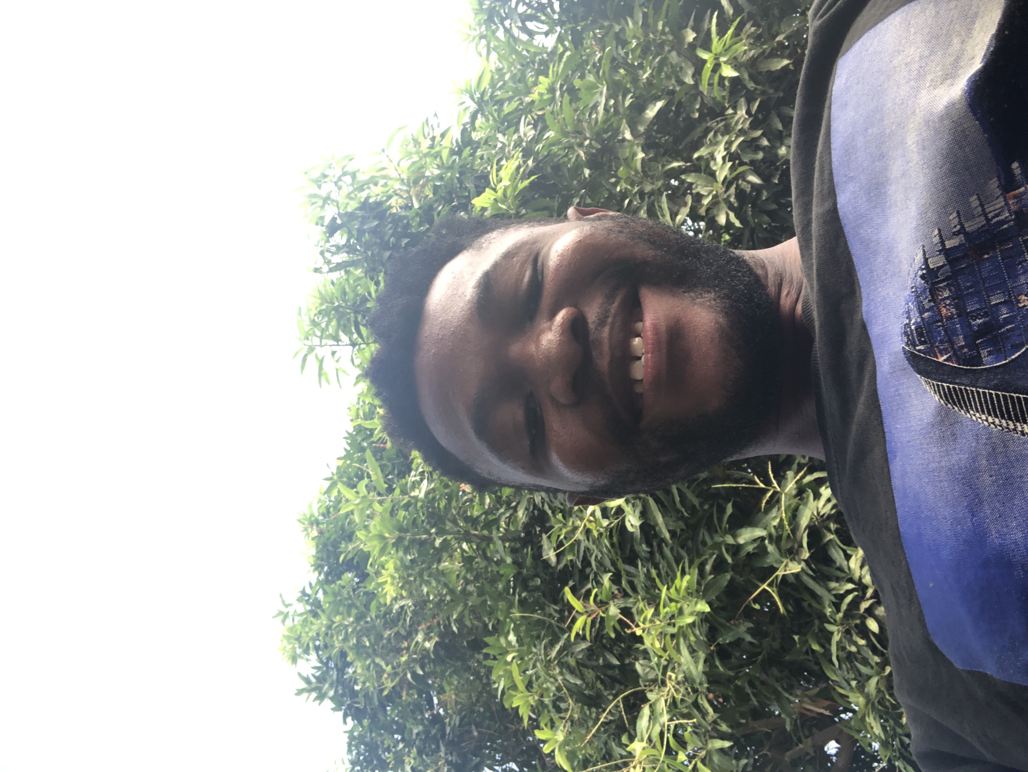 The image shows a person with dark skin, a beard, and short hair, smiling towards the camera. The person is wearing a black top with a blue and grey design on the right side. In the background, there is lush green foliage, possibly trees. Above the trees, a bright, overcast sky is visible. The location is Unknown, Unknown.