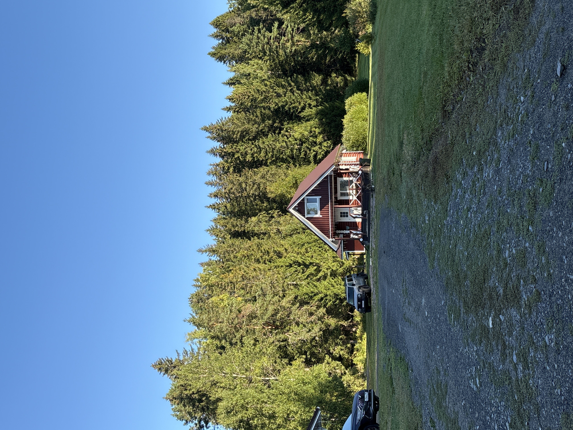 In the city of Rautalakhti, Russia, a picturesque scene unfolds under a clear blue sky. A rustic red wooden house with white trim sits nestled in a clearing, its gable roof a warm contrast to the lush green of the surrounding coniferous forest. A gravel driveway leads towards the house, where two vehicles are parked: a dark SUV on the left and a light-colored SUV to the right, closer to the house. The house features a small porch with white railings and a welcoming entrance. The forest behind the house is dense and tall, suggesting a natural, remote setting. The ground in front of the house is a mix of short, well-maintained grass and the gravel driveway, with some patches of taller vegetation visible towards the edges of the scene. The lighting suggests it is either early morning or late afternoon, with long shadows cast by the trees and the house.
