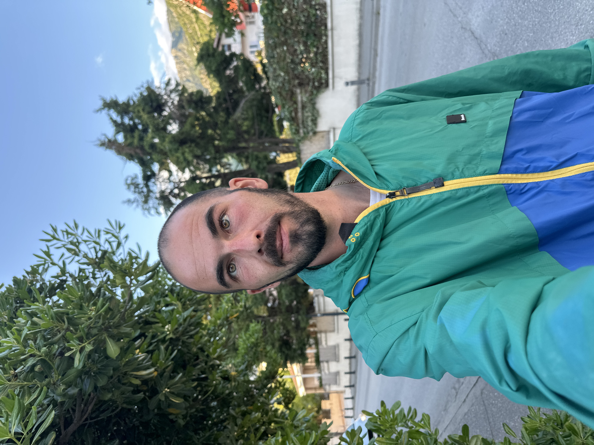 This is an outdoor selfie featuring a man, likely in his late 20s or 30s, with short dark hair, a trimmed beard, and prominent light green/hazel eyes. He looks directly at the camera with a neutral expression. He is wearing a striking multi-colored windbreaker jacket, featuring a vibrant green on the shoulders and arms, and a bold blue section across the chest and stomach. A bright yellow zipper runs down the center of the jacket, providing a strong contrast. A small black rectangular tag with a white number '1' is visible on the green part of his jacket, near his left shoulder. A silver chain is also visible around his neck.
The background reveals a sunny daytime scene with a clear blue sky and some scattered wispy clouds. Lush green foliage and dense trees are abundant, suggesting a vibrant natural environment. In the mid-ground and distance, residential buildings with light-colored facades and reddish-orange roofs are visible, along with the silhouette of mountains under the clear sky. The overall impression is that of a pleasant day in a scenic location. Given the context, the setting is Budva, Montenegro, known for its mountainous terrain, coastal views, and Mediterranean architecture. The bright lighting indicates late morning or early afternoon.