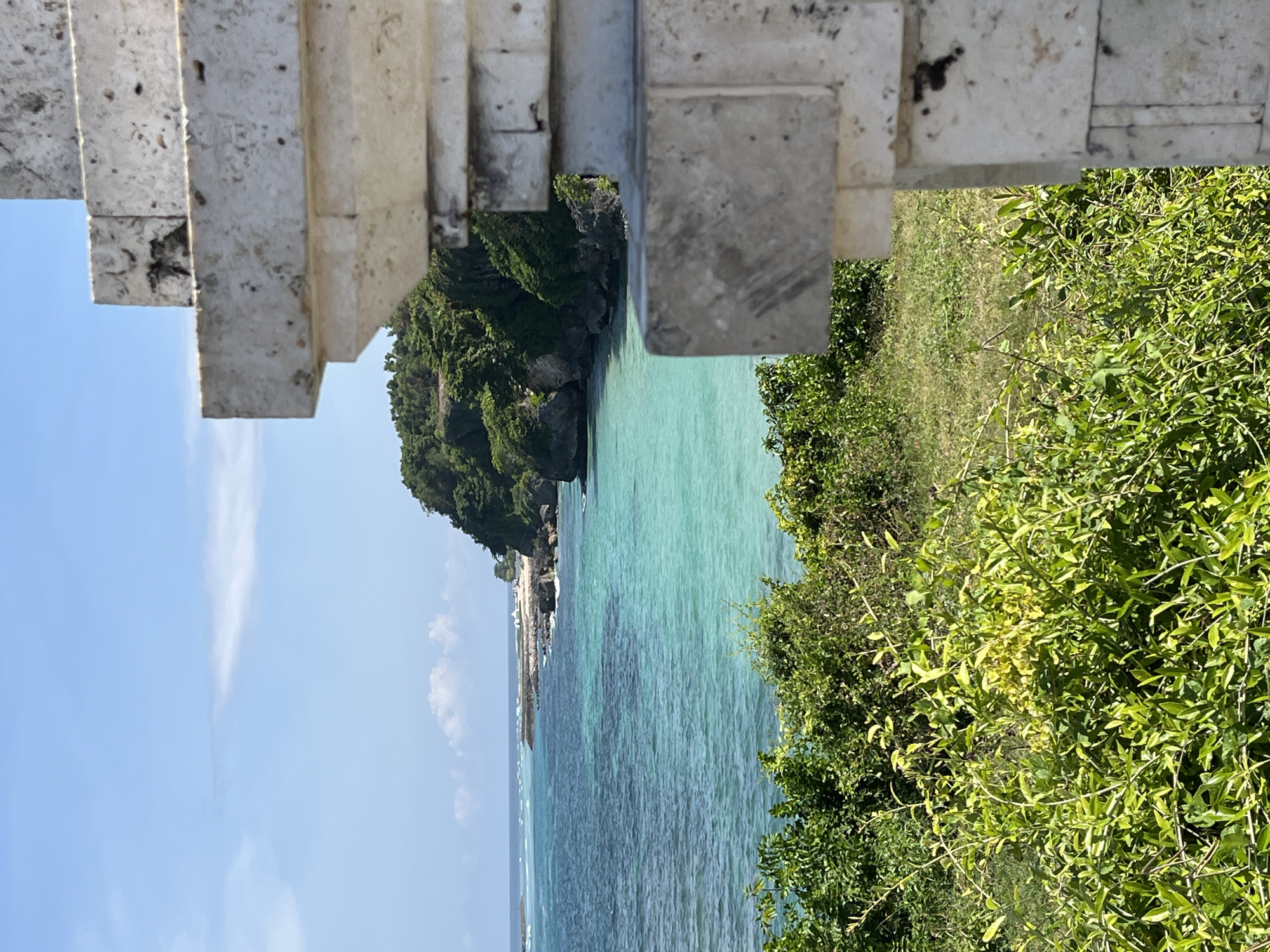 The image captures a serene, sun-drenched coastal landscape, likely taken from an elevated vantage point. In the foreground, a robust stone structure, possibly a set of steps or a decorative wall, frames the upper and right portions of the scene. The stone is light grey, textured, and shows signs of weathering with visible grains and some dark spots.
Lush, vibrant green foliage, comprising various bushes and trees, dominates the lower right corner and extends into the midground, covering distant cliffs. The core of the image is an expansive view of the ocean, which displays a stunning gradient of colors. Near the shore, the water is a brilliant turquoise, indicative of clarity and shallow depths, gradually deepening to richer shades of blue further out. Darker patches beneath the surface suggest underwater features like coral reefs or deeper sections.
Along the distant left side of the frame, verdant, tree-covered cliffs rise majestically from the water, with a small, light-colored strip of sand or a beach visible at their base. The background features a clear, expansive light blue sky, accented by a few faint, wispy white clouds.
The image depicts a bright, clear day, likely midday or early afternoon, given the strong illumination and lack of long shadows. There are no people visible, and no discernible text. The scene evokes a sense of natural beauty and tranquility, consistent with a tropical location such as Kuta Selatan, Indonesia.