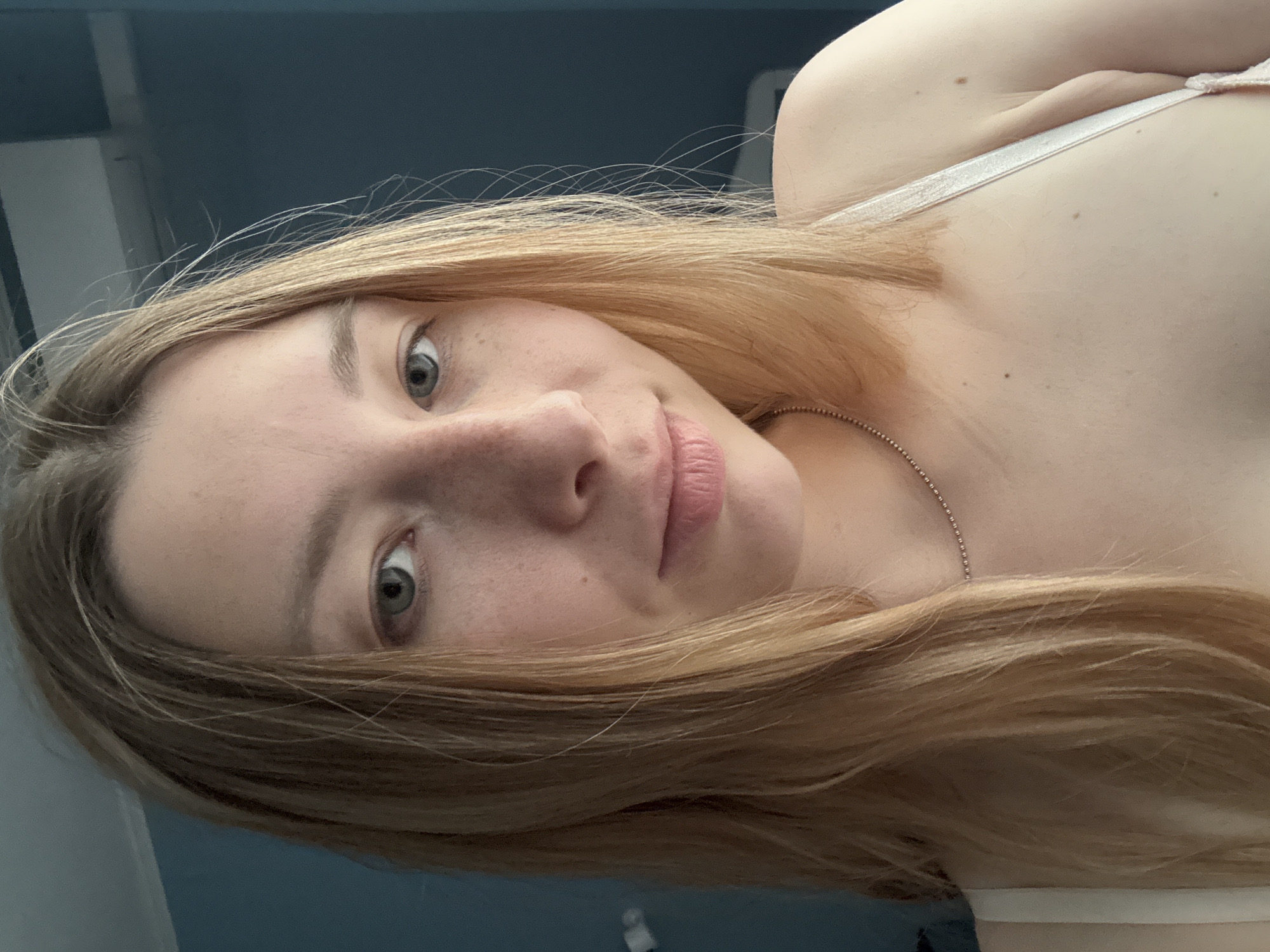 A close-up, slightly angled portrait of a fair-skinned woman with light brown to blonde hair. Her hair is parted to the side, falling around her face and shoulder. She has freckles on her face, particularly around her nose. Her eyes are light colored, possibly green or hazel. She is wearing a light-colored tank top with thin straps and a silver necklace. The background is blurry and includes a blue wall and a white door or frame. There is no indication of location in Moscow, Russia.