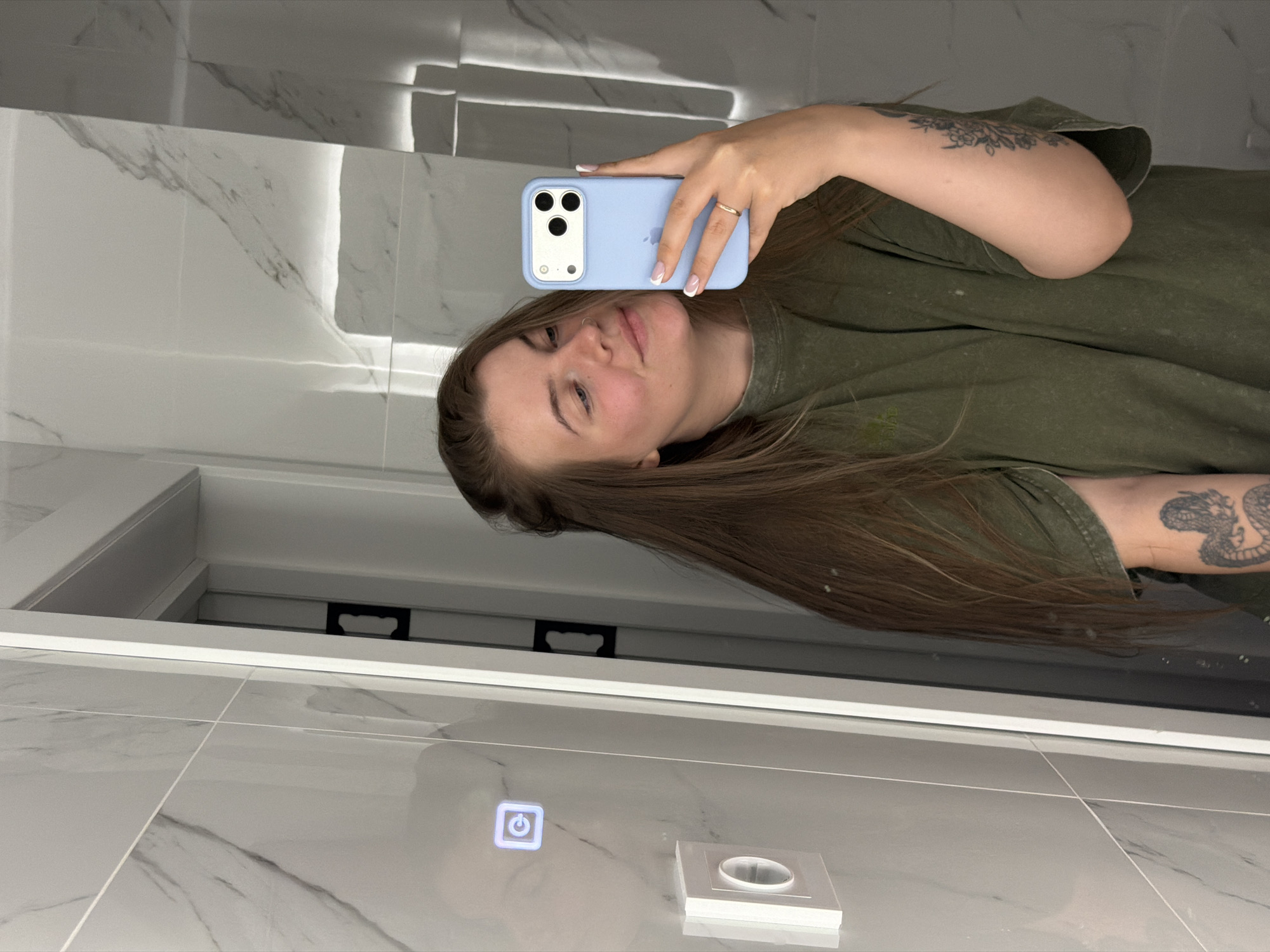 A woman takes a selfie in a bathroom. She holds a light blue iPhone in her right hand. She has long brown hair, and is wearing an olive green t-shirt. She has tattoos on her right arm. The bathroom has gray marble tiles and a lighted mirror. There is a light switch on the wall. Based on the user's location data, it's likely the image was taken in Chaikovsky, Russia.