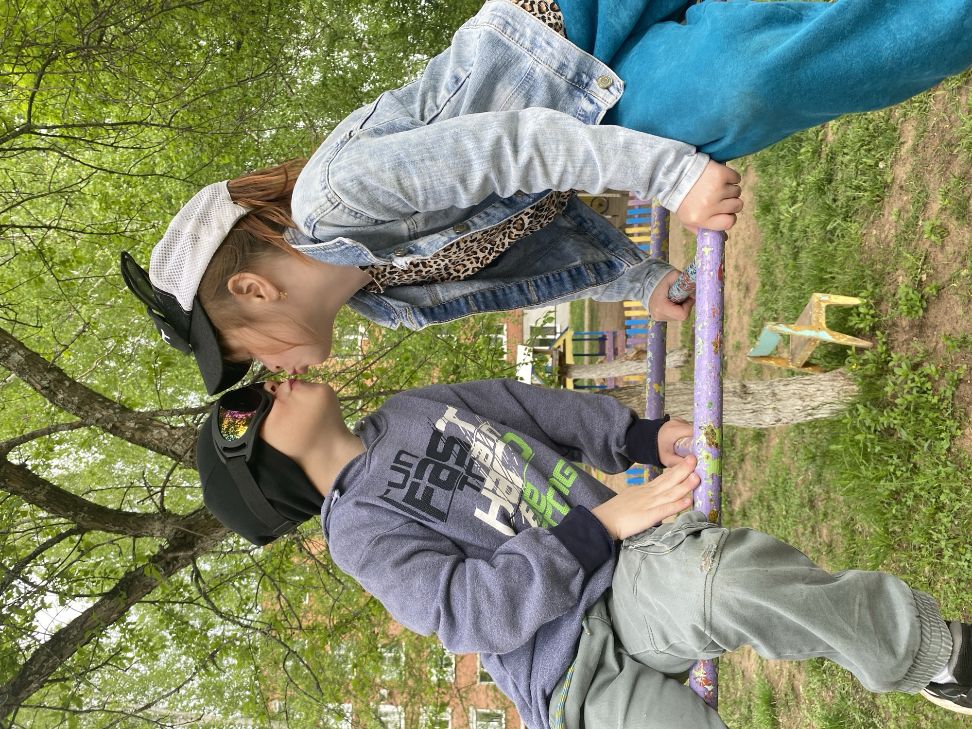 Two children are on a piece of playground equipment painted purple. One child wears a denim jacket, a leopard print shirt, blue pants, and a black and white baseball cap. The other child wears a grey sweatshirt, green pants, a black baseball cap, and iridescent goggles. They appear to be in a park. A building is visible in the background, suggesting an urban environment. The location is Noviy, Russia.