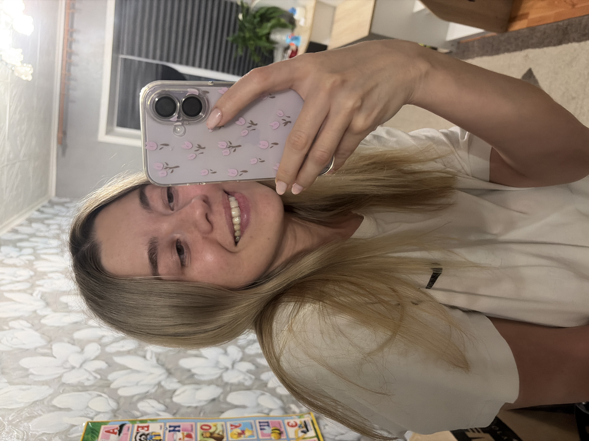 A woman is taking a selfie with a phone case decorated with pink flowers. She has blonde hair, light skin, and is wearing a white shirt. The background includes a room with floral wallpaper, a window with blinds, and a plant. Part of an alphabet learning toy is visible in the foreground. Based on other image results, this photo could have been taken in Chaikovsky, Russia.