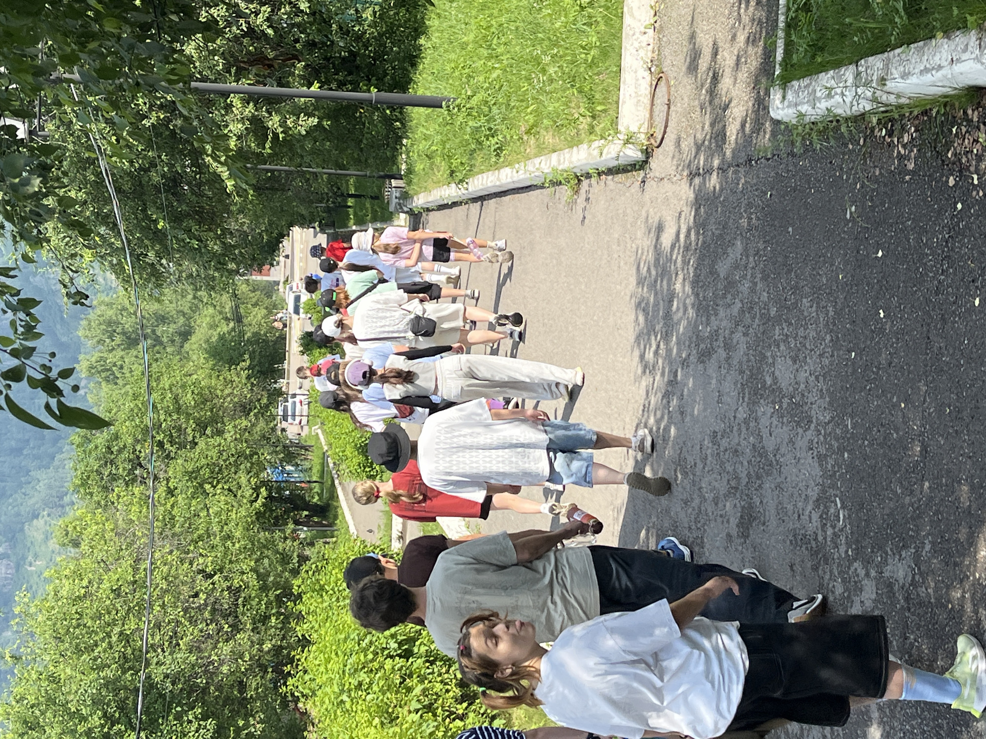 A group of approximately 15 people, appearing to be a mix of children and adults, are walking single file along a paved pathway in Divnogorsk, Russia. The pathway is bordered by lush green vegetation and trees. In the background, a mountainous landscape is visible, partially obscured by trees. The sky is clear and bright, indicating a sunny day. The individuals are dressed in casual summer clothing. Some are holding water bottles. The pathway appears to be on a slight incline.